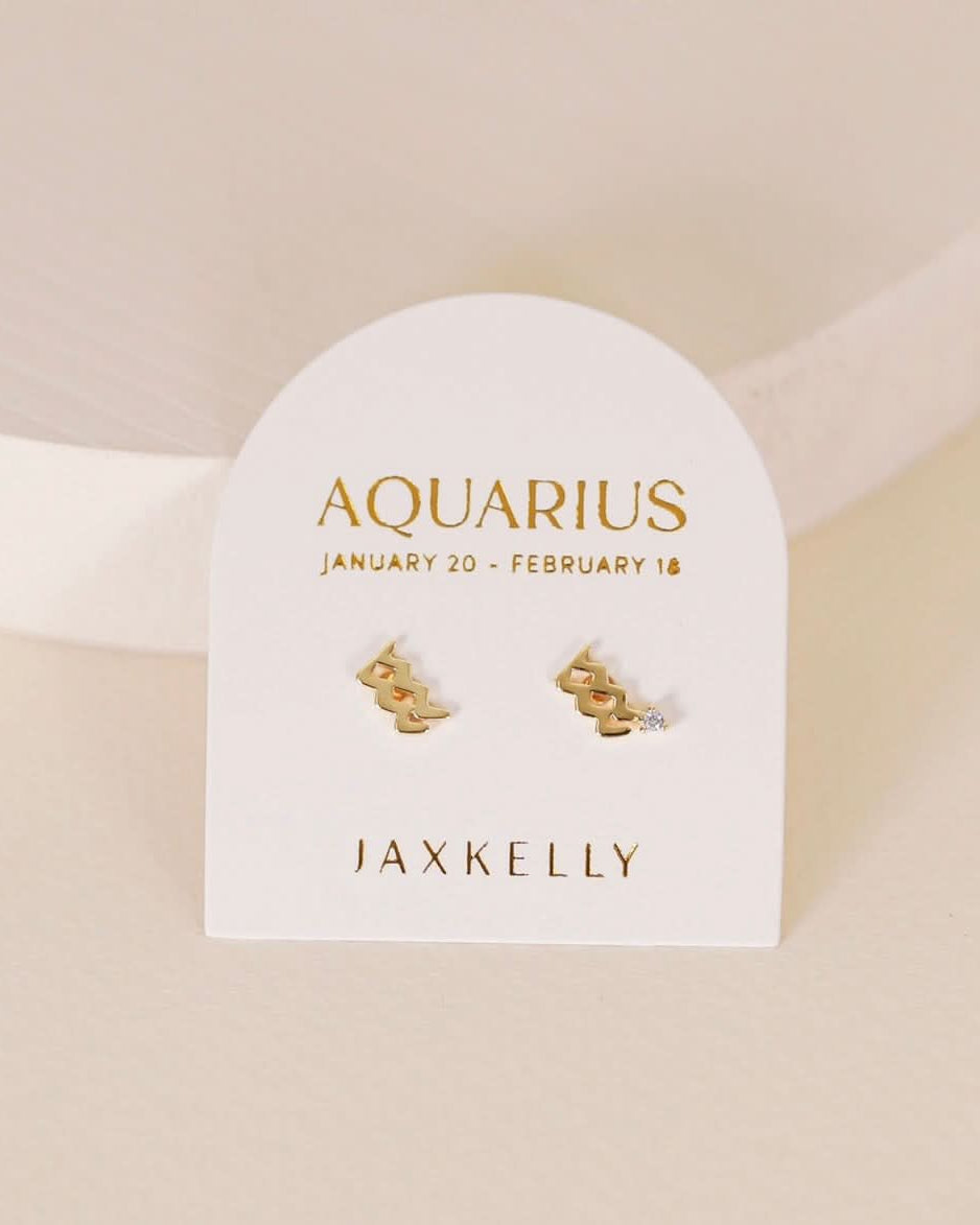 Gold zodiac charm studs in the shape of Aquarius astrological sign displayed on earring card