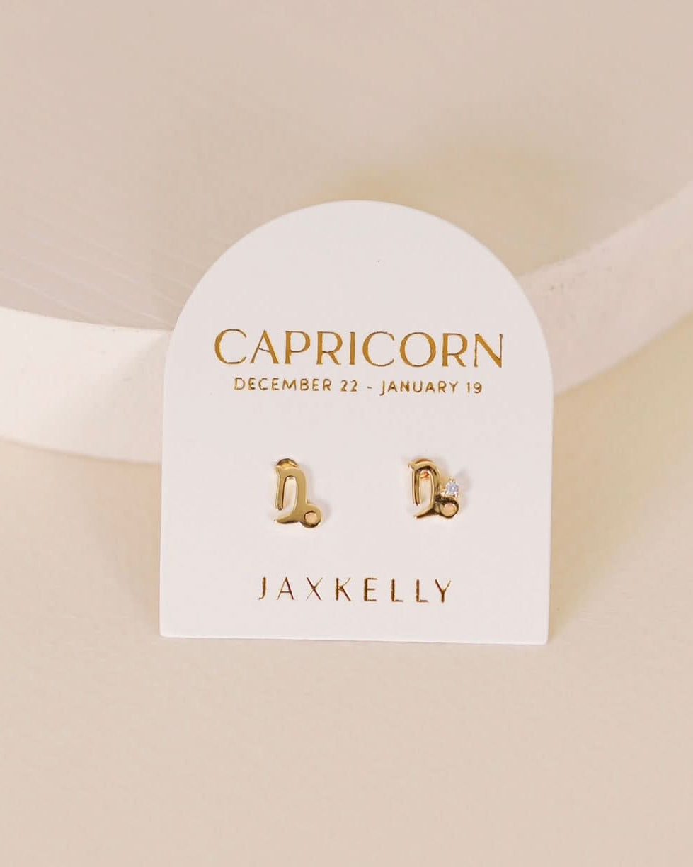 Gold zodiac charm studs in the shape of Capricorn astrological sign displayed on earring card