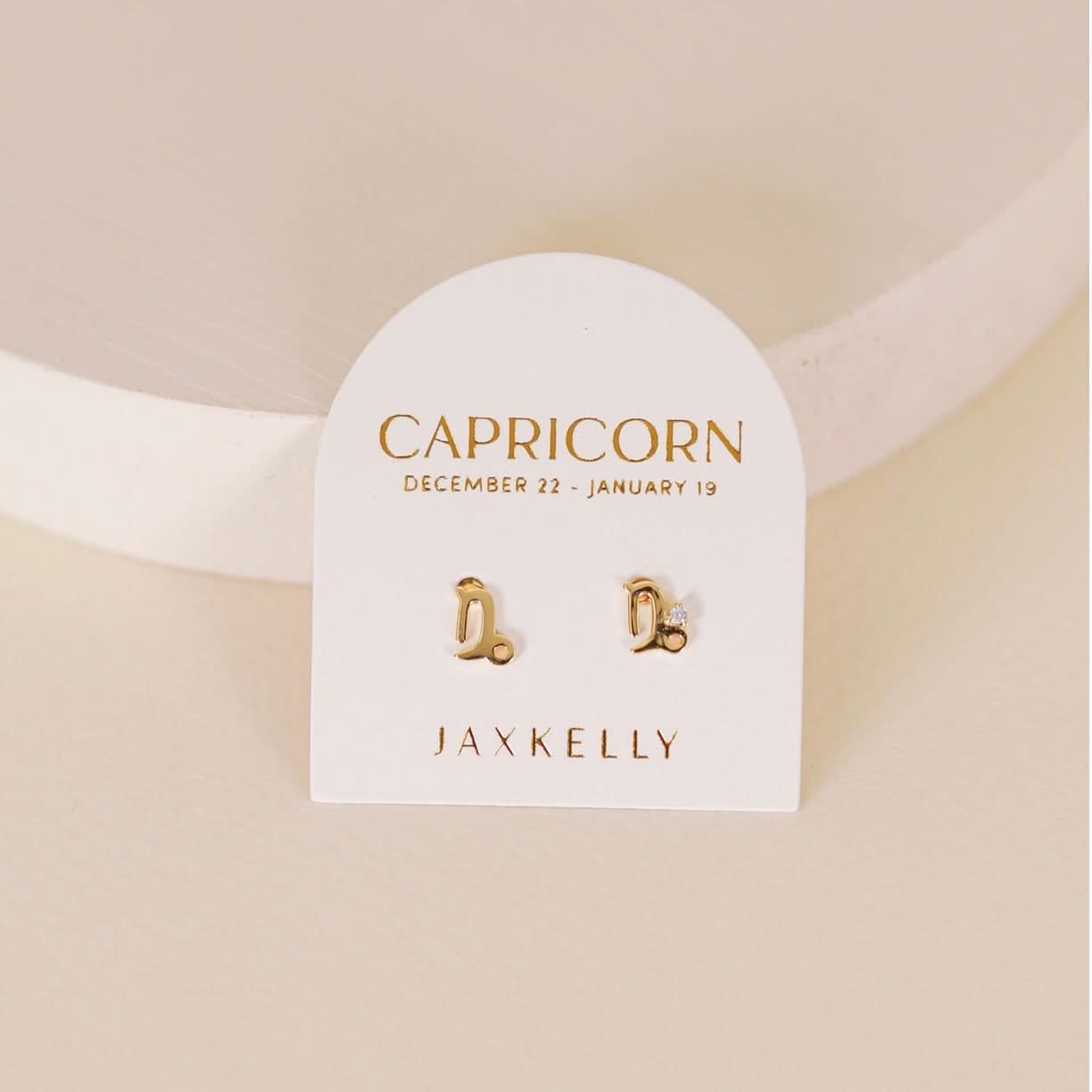 Gold zodiac charm studs in the shape of Capricorn astrological sign displayed on earring card