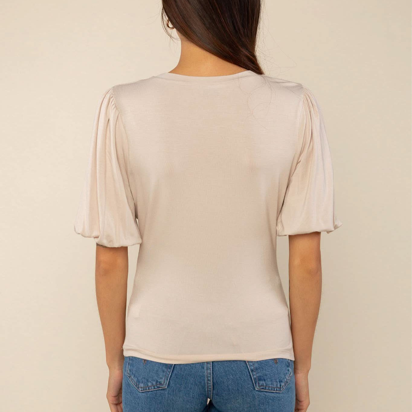 Woman wearing a sand color crew neck top with puffy sleeves and blue jeans with back pockets behind a blank background