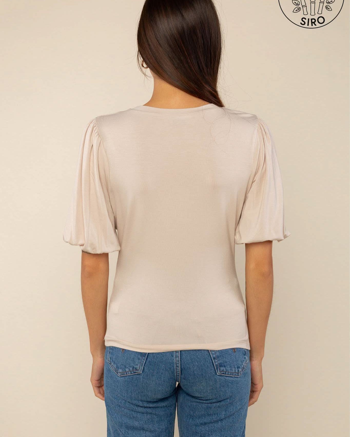 Woman wearing a sand color crew neck top with puffy sleeves and blue jeans with back pockets behind a blank background