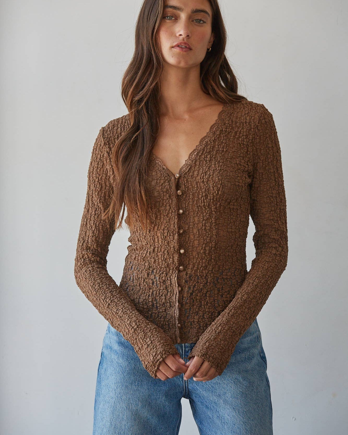 A woman wearing a brown textured knit cardigan with a scalloped V-neck and button-front closure, paired with light blue jeans, photographed against a plain light gray studio background.
