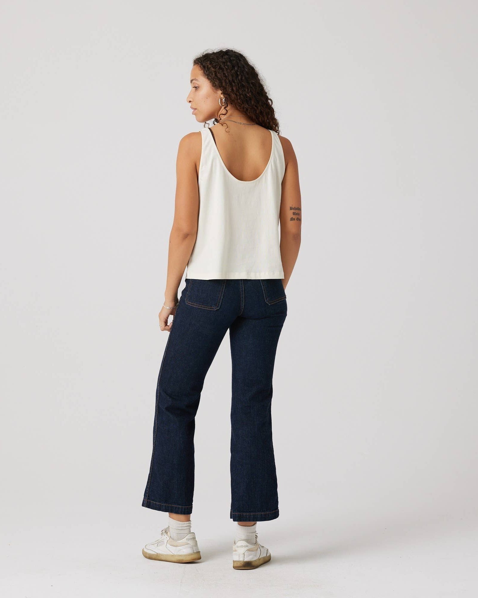 Person wearing a white tank top and dark blue jeans on a plain background