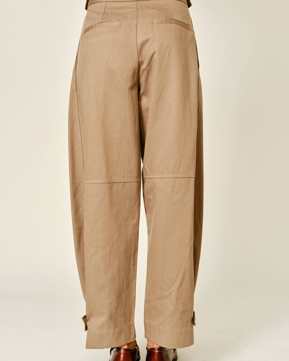 A person shown from the back wearing loose, wide-leg tan pants with belt loops, flap back pockets, and cuffed hems, paired with brown shoes against a plain light studio background.