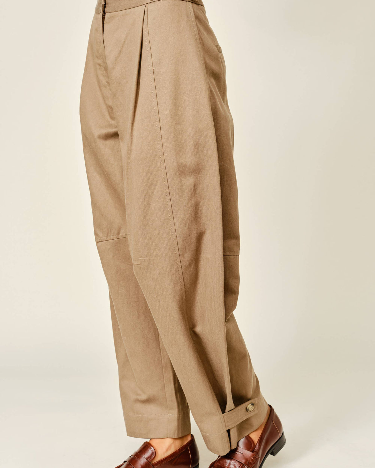A person wearing high-waisted, wide-leg pleated trousers in a warm khaki-beige color, paired with brown leather loafers, shown from the waist down against a plain light beige studio background.