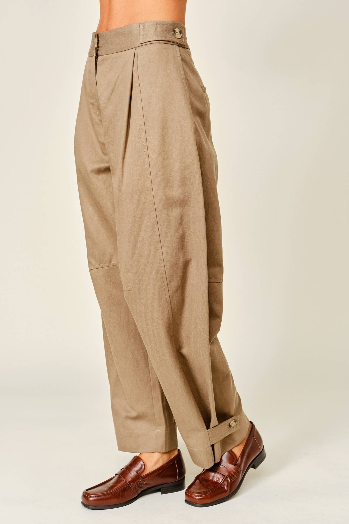 A person wearing high-waisted, wide-leg pleated trousers in a warm khaki-beige color, paired with brown leather loafers, shown from the waist down against a plain light beige studio background.