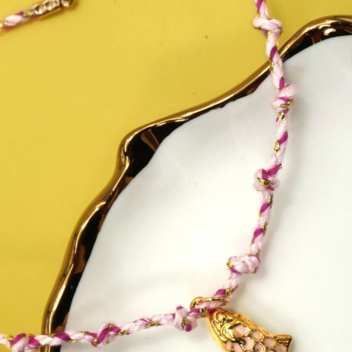 Pink braided bracelet with a gold fish charm on a white dish against a yellow background