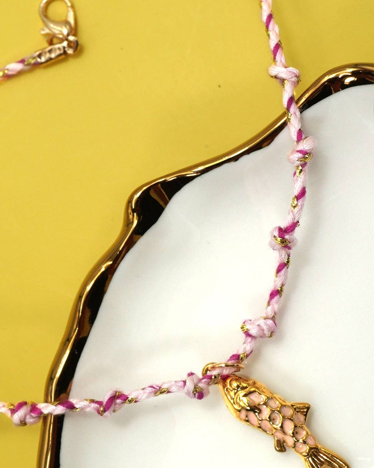 Pink braided bracelet with a gold fish charm on a white dish against a yellow background