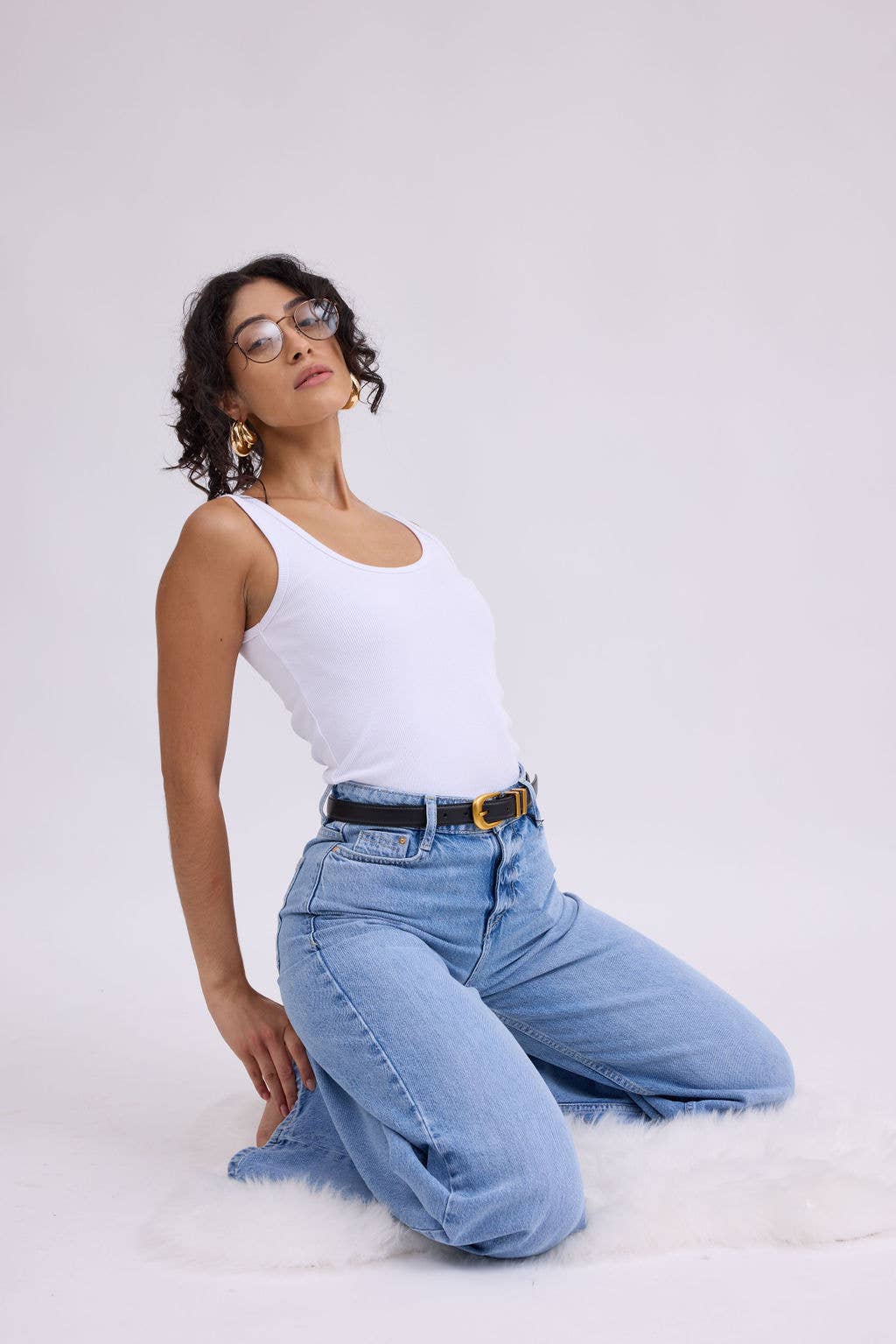 Woman wearing a white tank top and blue jeans sitting on a white background