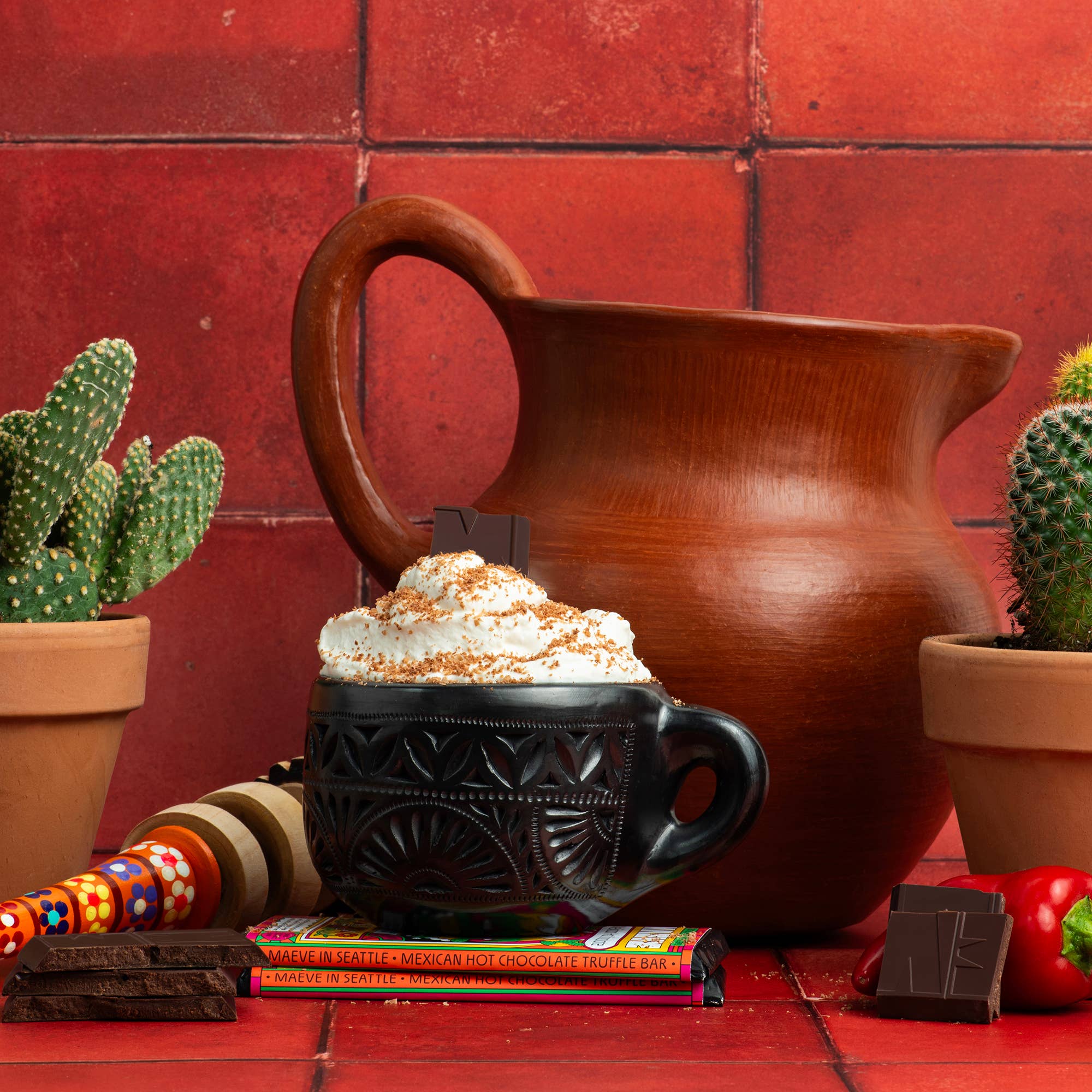 Hot chocolate with whipped cream in a black mug, surrounded by a brown pitcher, cacti, and candy on a red tiled background.