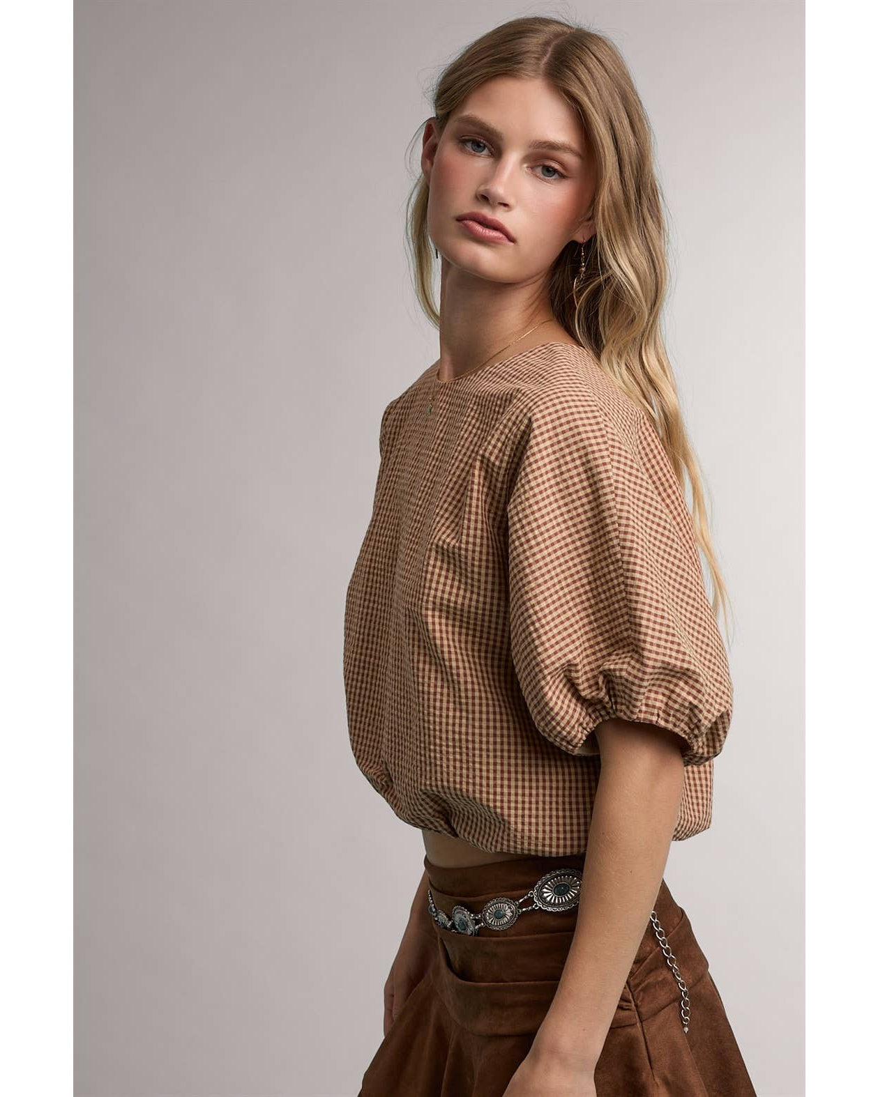 A woman modeling a brown and beige checkered puff-sleeve top with a cropped silhouette, paired with a brown skirt and a silver chain belt, against a plain gray studio background.