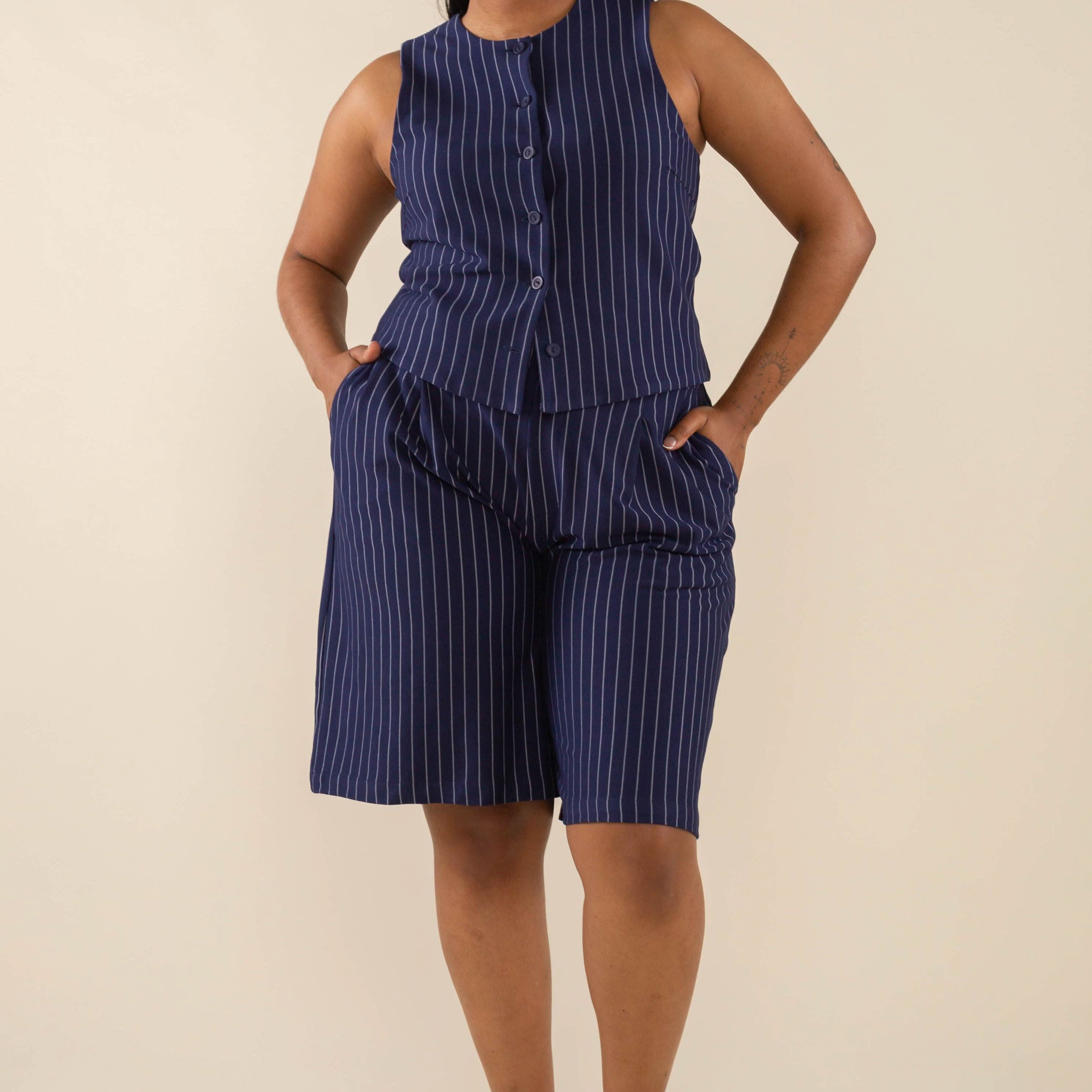 Woman wearing a navy vertical pin striped button down sleeveless top with navy vertical pin striped knee length shorts with pockets behind a blank background.