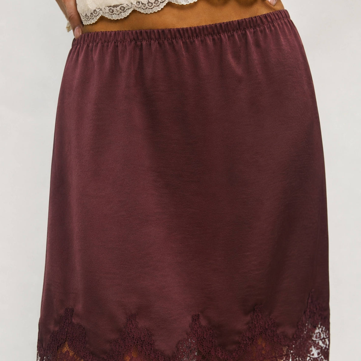 A close-up of a model wearing a dark burgundy slip skirt with a lace hem and a matching lace waistband.