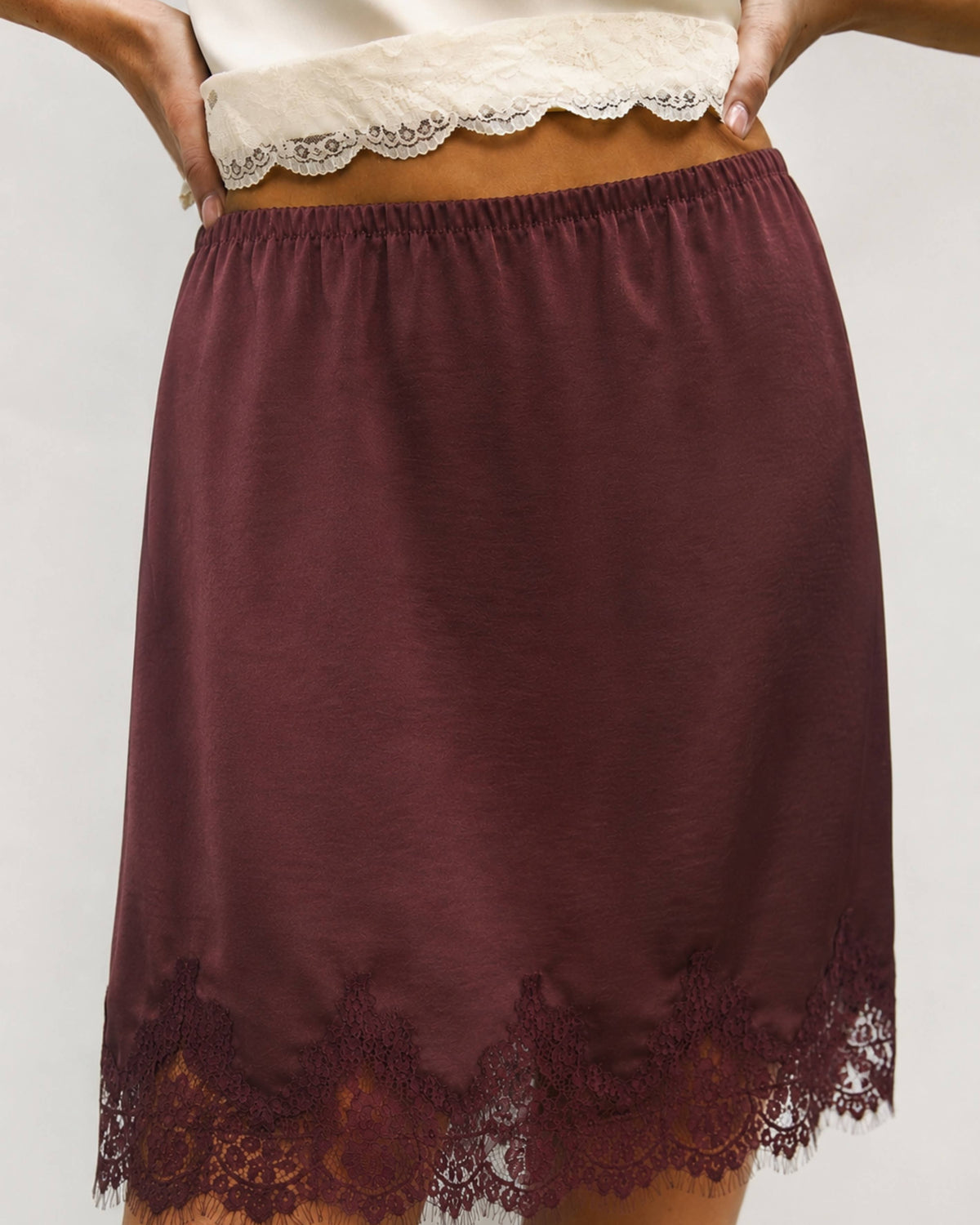 A close-up of a model wearing a dark burgundy slip skirt with a lace hem and a matching lace waistband.