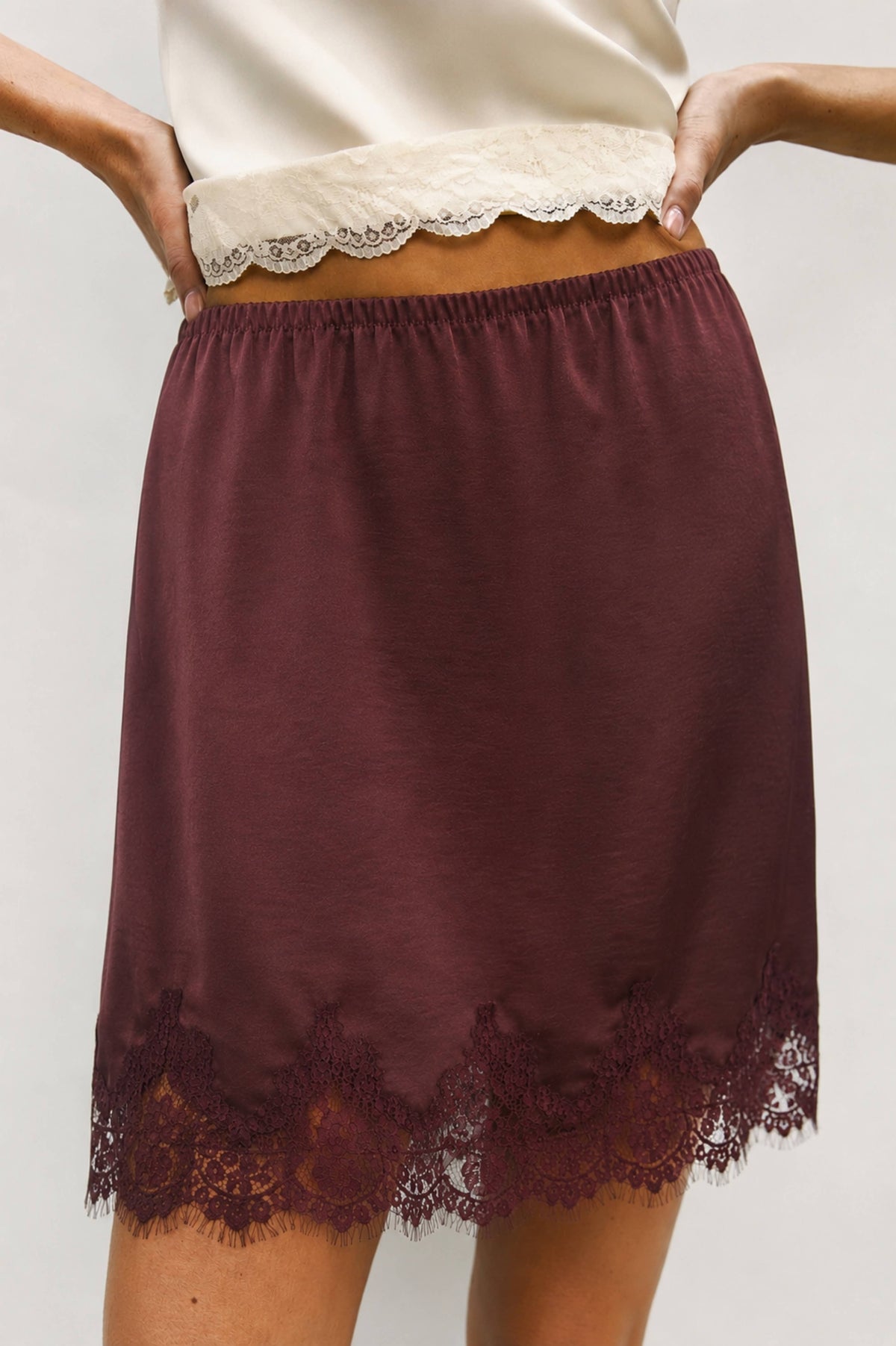 A close-up of a model wearing a dark burgundy slip skirt with a lace hem and a matching lace waistband.