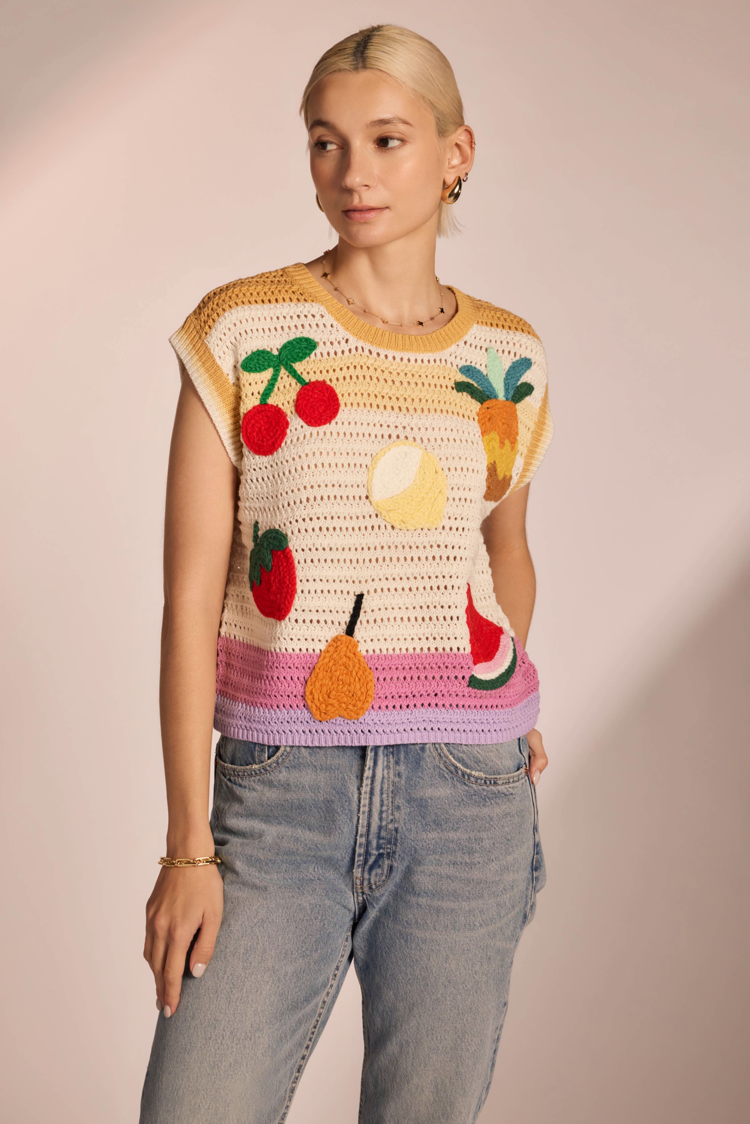 Woman wearing sleeveless, ivory, colorful, crochet, top with cherries, lemon, strawberry, watermelon, pear, and pineapple with pink, purple, and yellow horizontal stripes