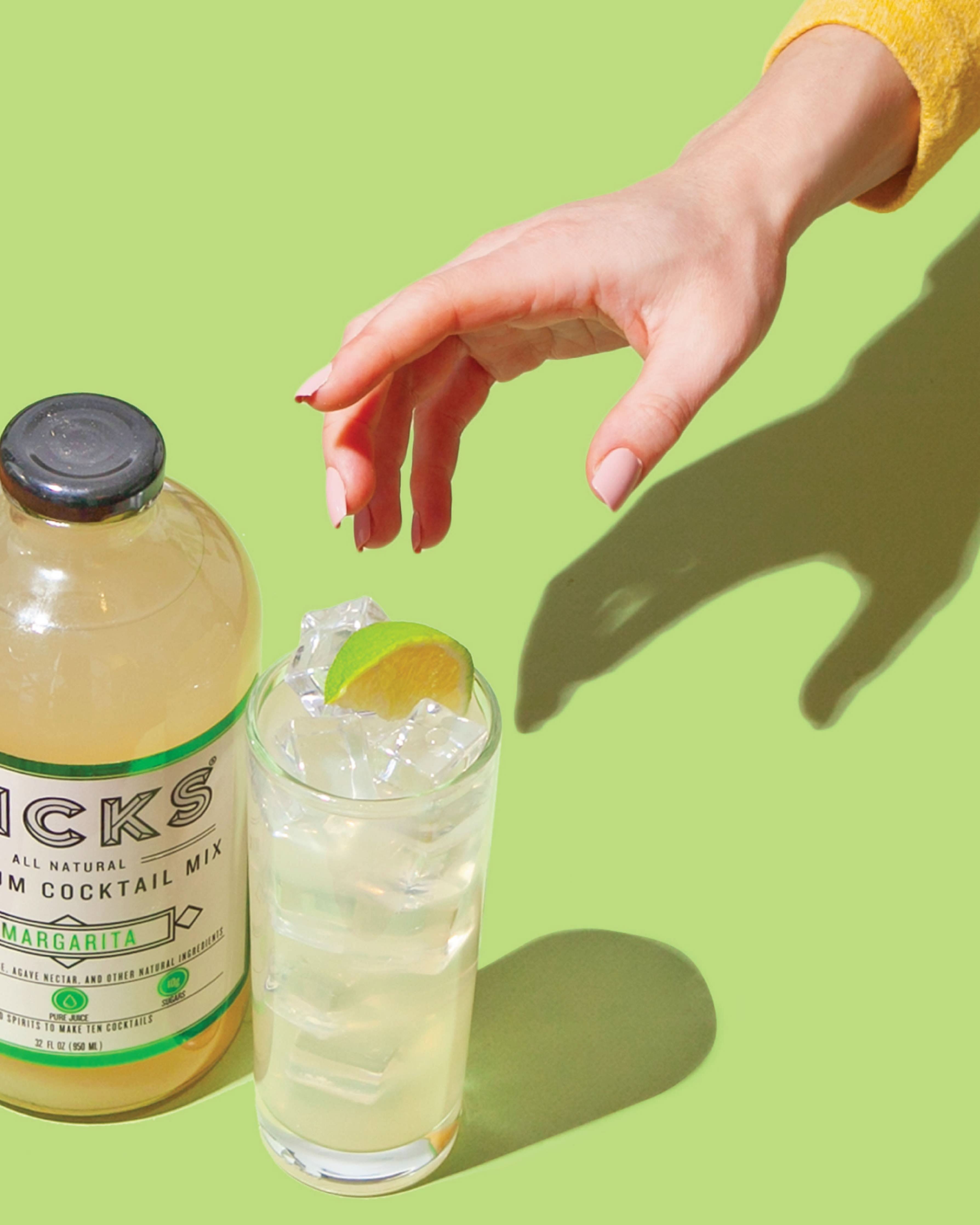 Glass cocktail bottle sitting on green surface next to drinking glass containing cocktail with hand reaching for glass