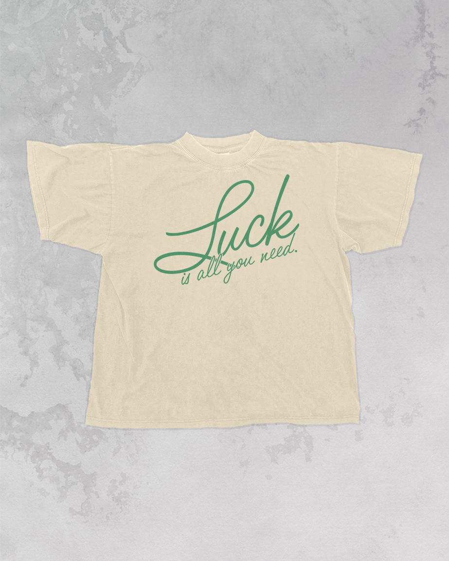 “Luck is all you need” written in green cursive on cream tee shirt playing on a gray marbled backgound