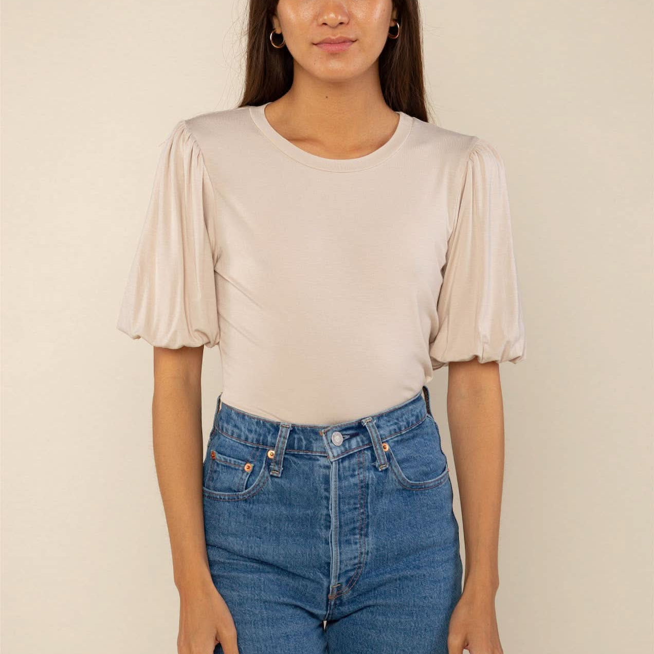 Woman wearing a sand color crew neck top with puffy sleeves and blue jeans behind a blank background.
