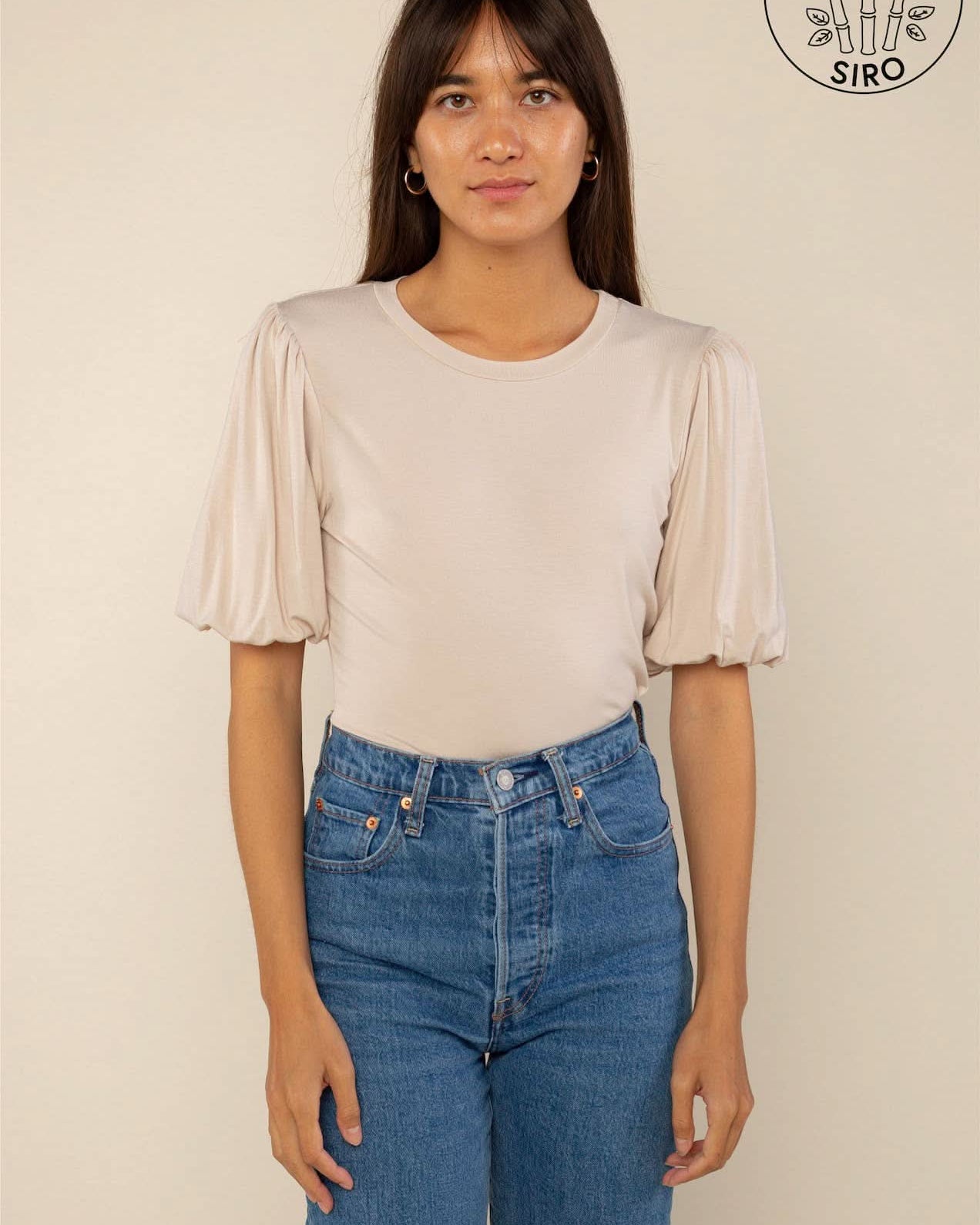 Woman wearing a sand color crew neck top with puffy sleeves and blue jeans behind a blank background.