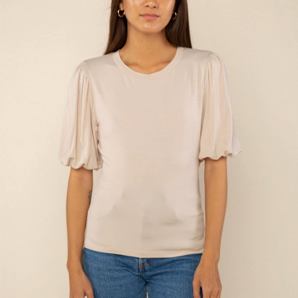 Woman wearing a sand color crew neck top with puffy sleeves and blue jeans behind a blank background