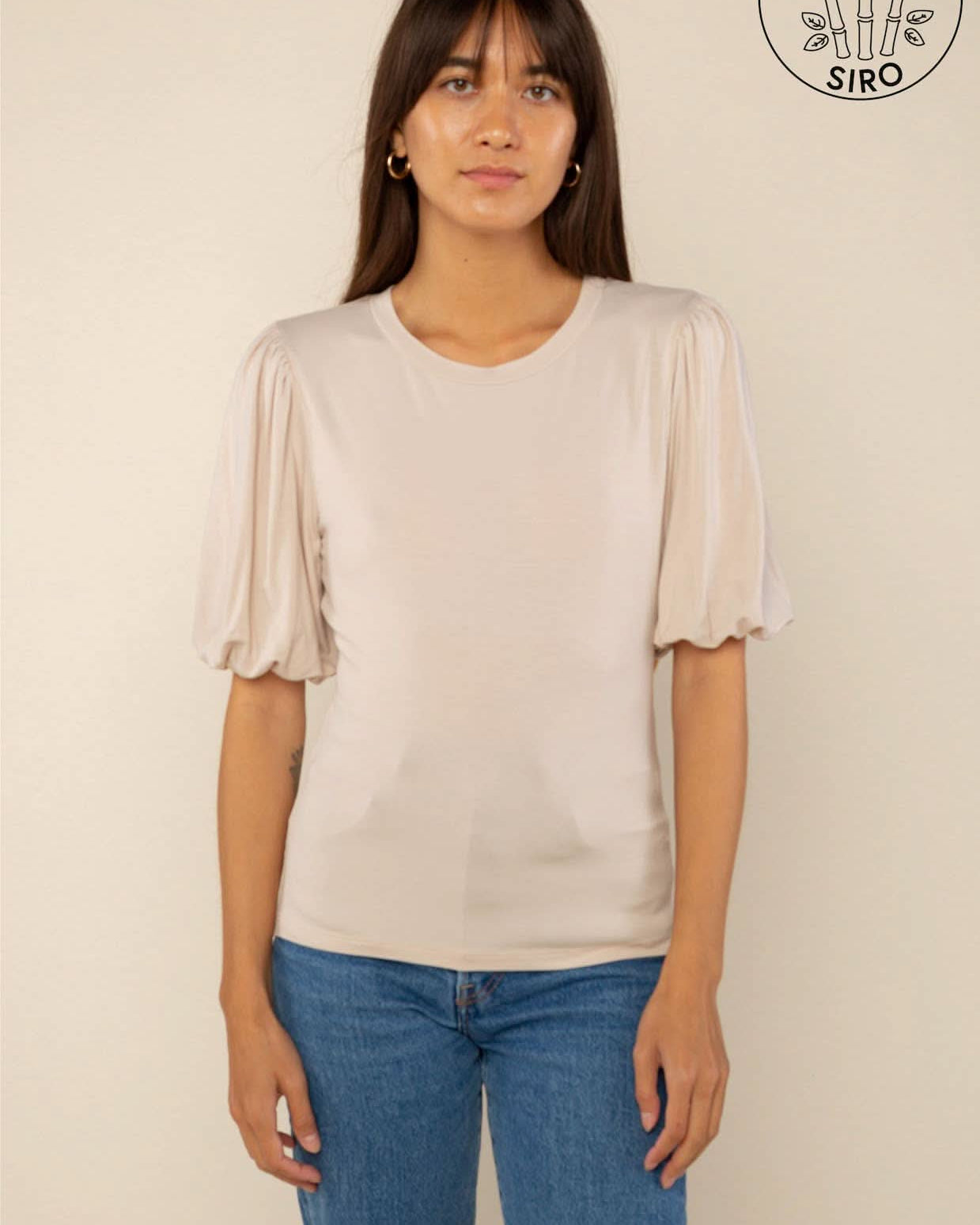 Woman wearing a sand color crew neck top with puffy sleeves and blue jeans behind a blank background