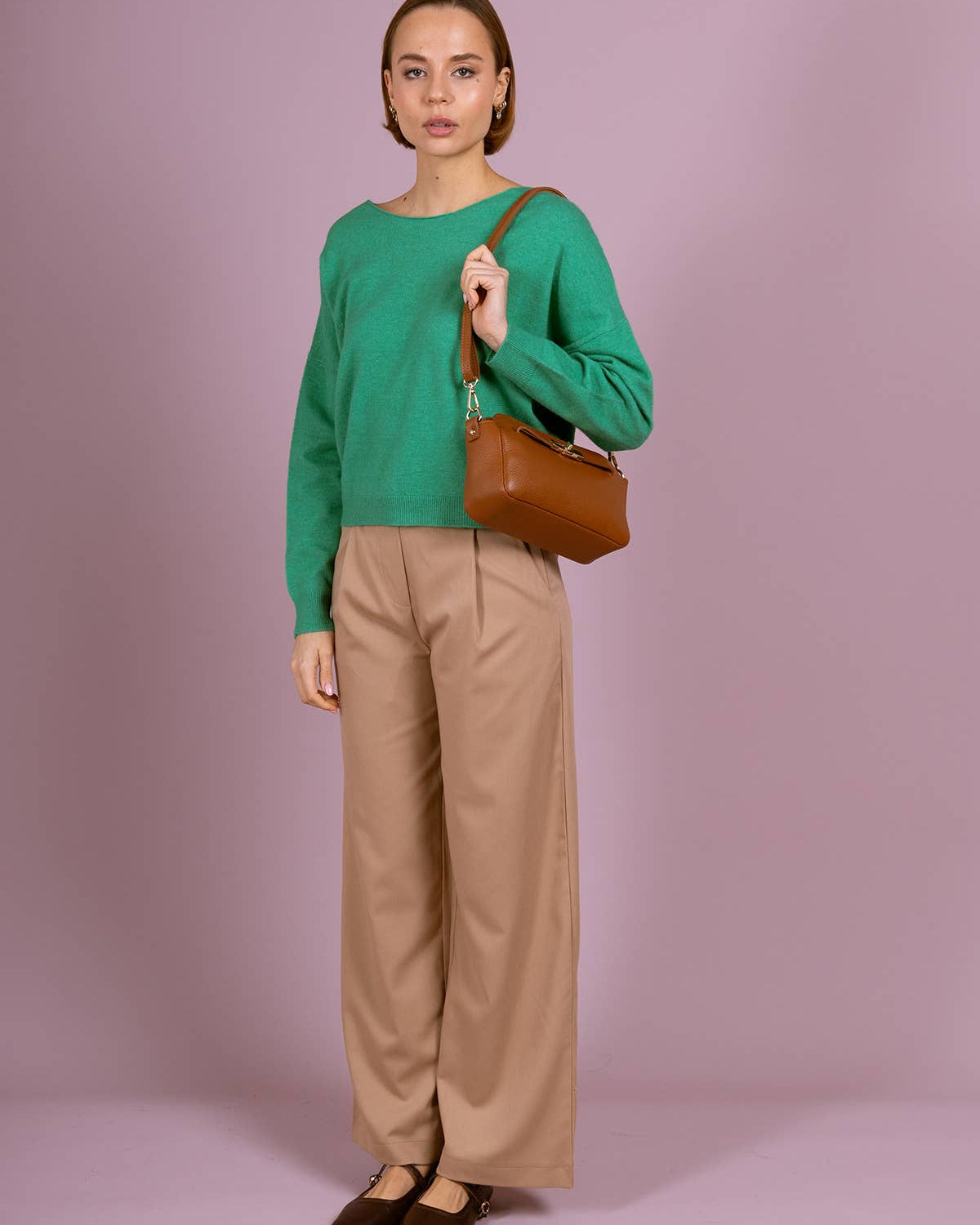 A woman stands against a solid lavender studio background wearing a vibrant green long-sleeve knit sweater, beige wide-leg trousers, and dark brown flats. She is holding a brown leather shoulder bag.