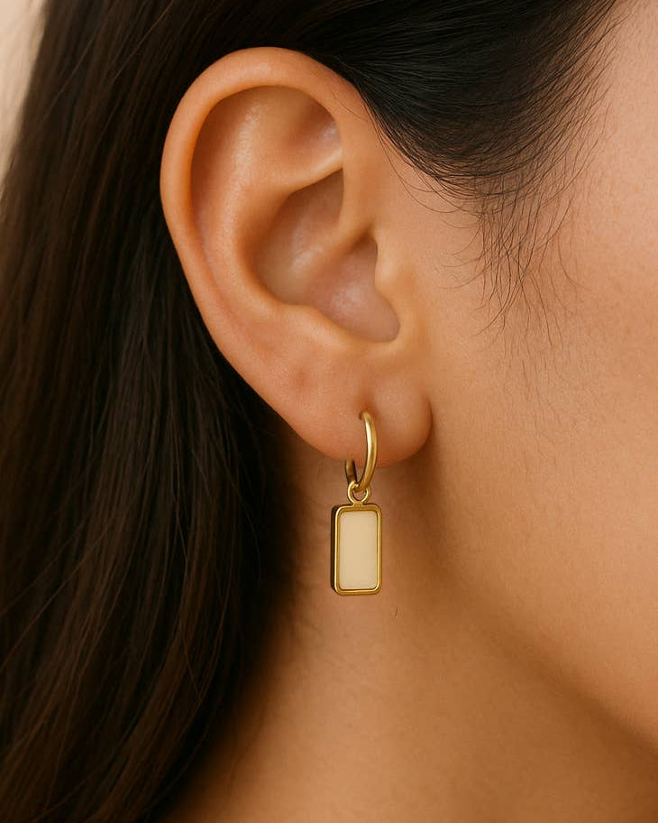 Gold hoop earring with a small rectangular pendant worn by a person.