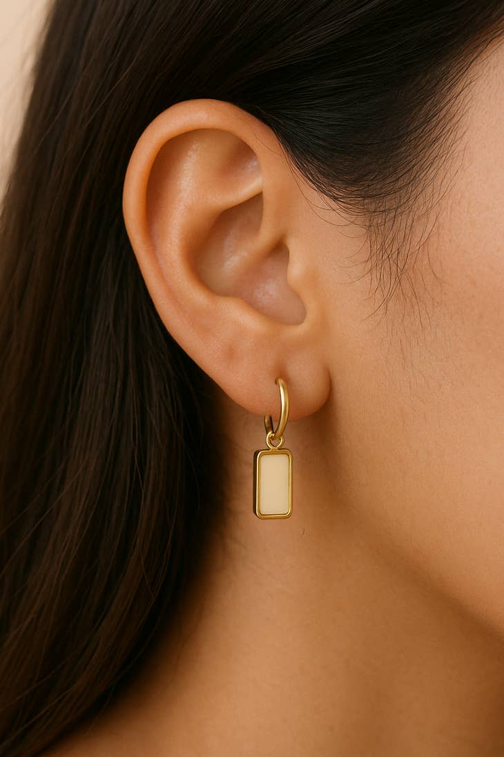 Gold hoop earring with a small rectangular pendant worn by a person.