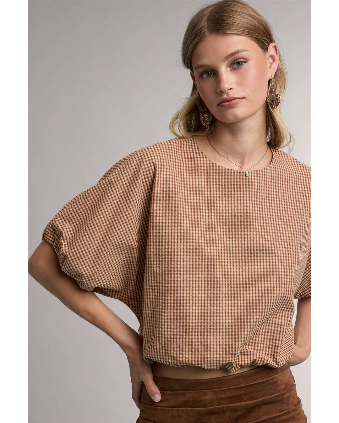 A woman wearing a brown and beige gingham short-sleeve top with voluminous puff sleeves, paired with a brown skirt, against a neutral gray studio background.