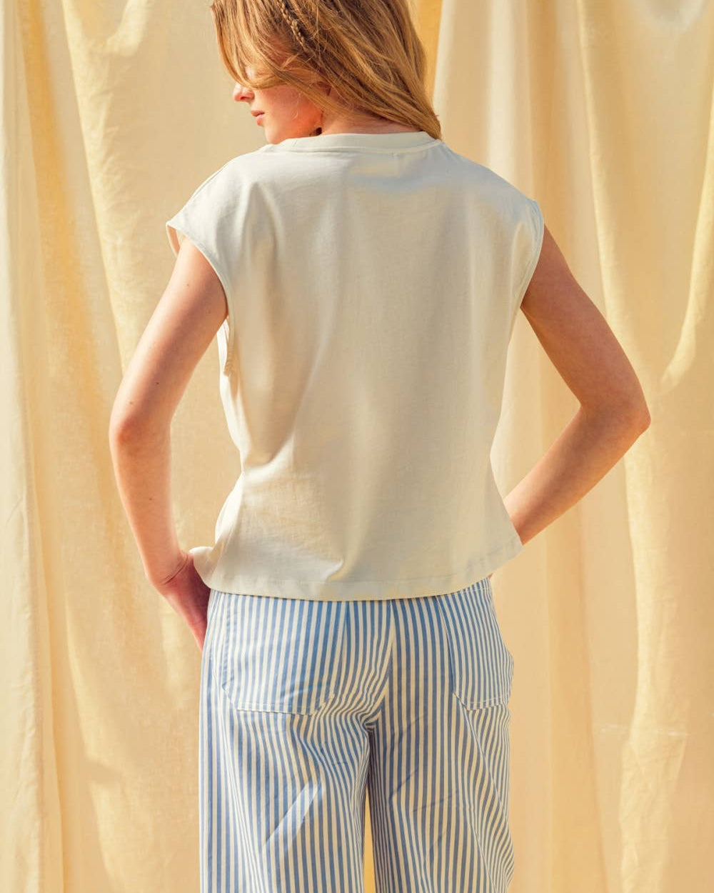 Person wearing a white sleeveless top and striped pants against a beige curtain.