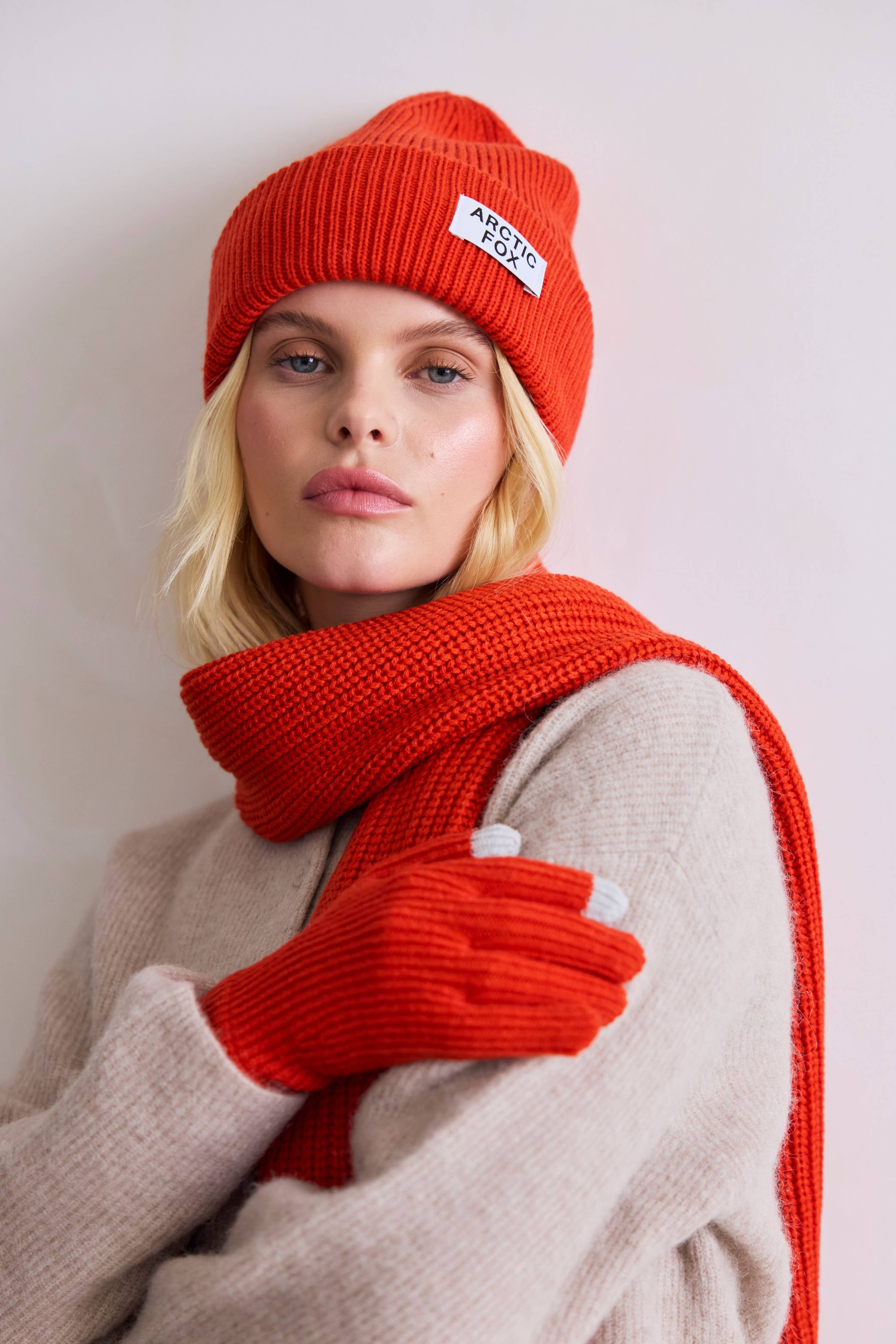 Person wearing a red knit hat, scarf, and gloves with a visible brand logo on a light background