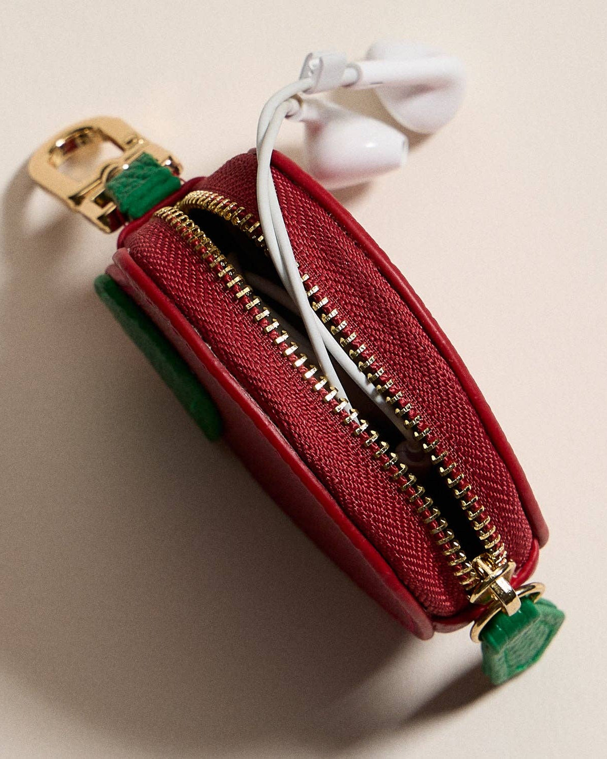 Red apple pouch with gold zipper and green accents containing white earbuds on a beige background