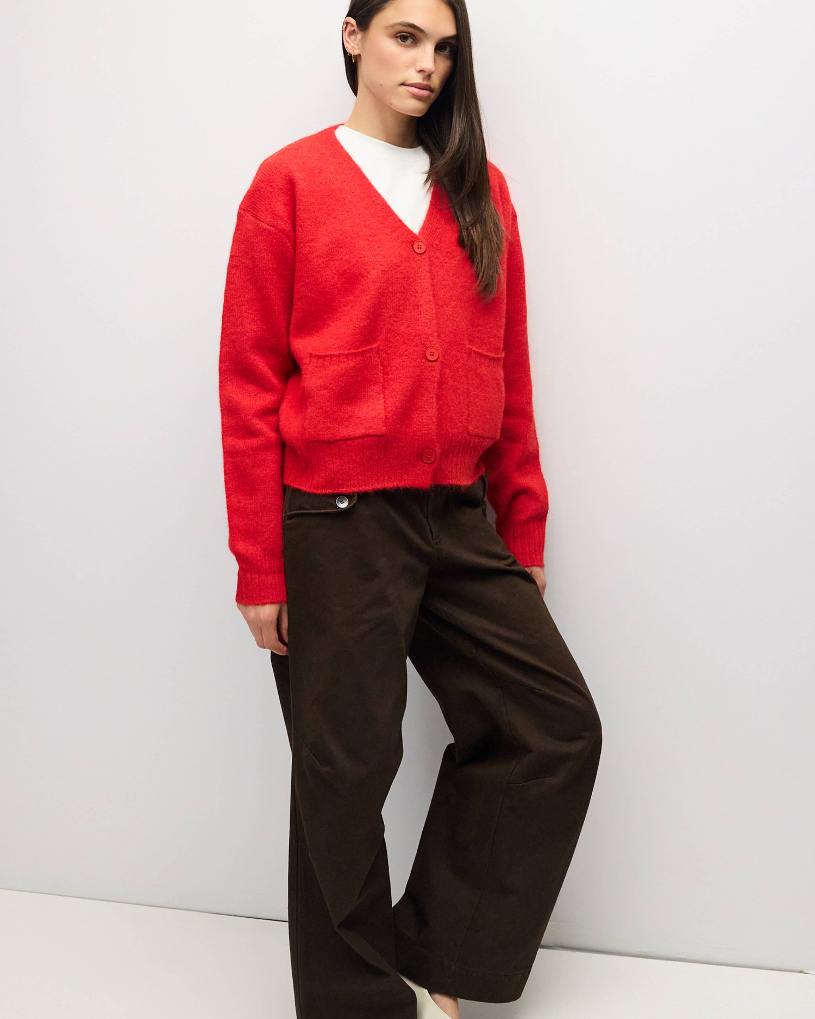 A woman modeling a bright red knit cardigan sweater with front buttons and patch pockets, paired with dark brown wide-leg trousers and white pointed flats, standing against a plain light gray studio wall.