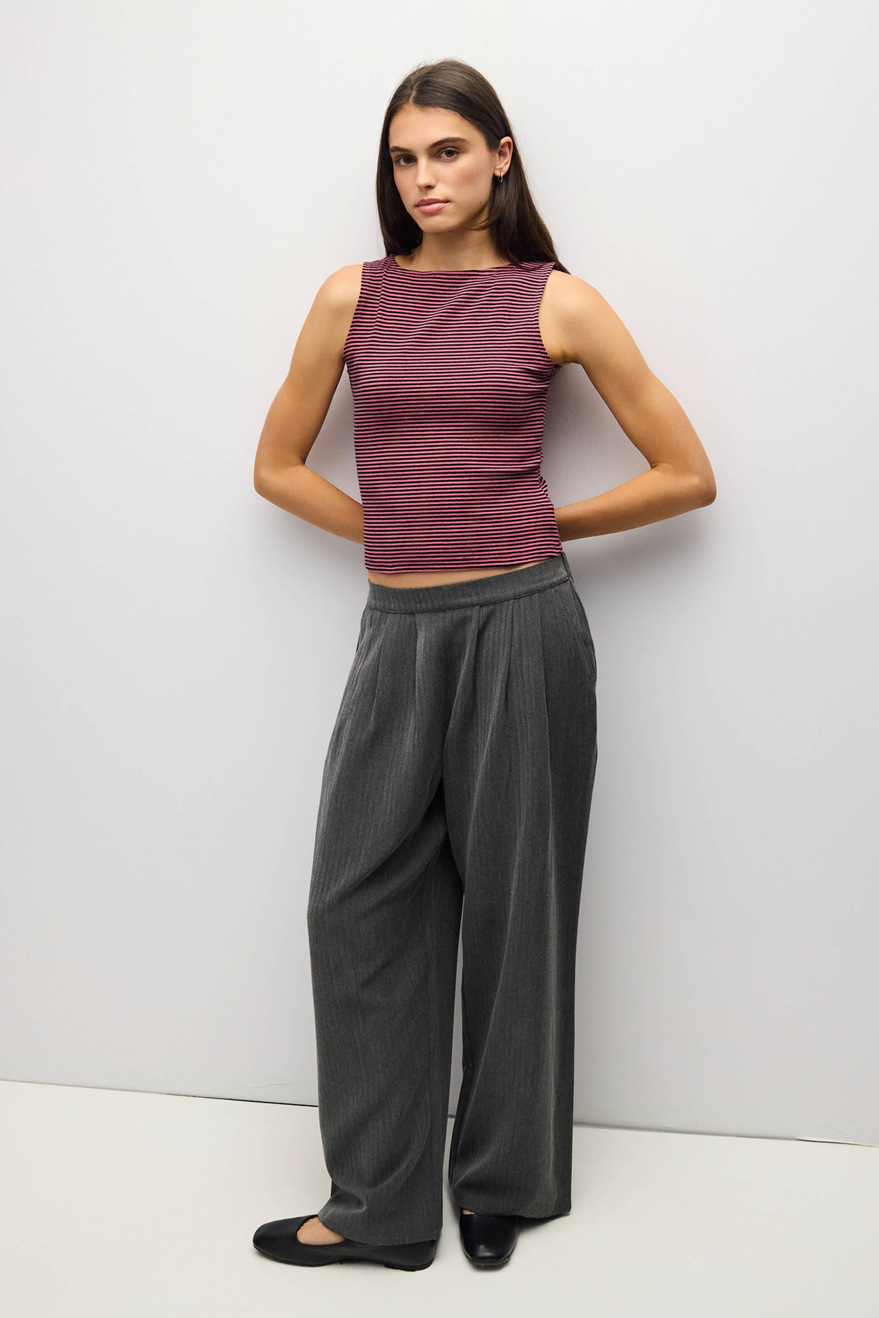 A woman modeling a sleeveless ribbed knit top in heathered burgundy and wide-leg pleated trousers in dark gray, standing against a plain light gray studio wall.