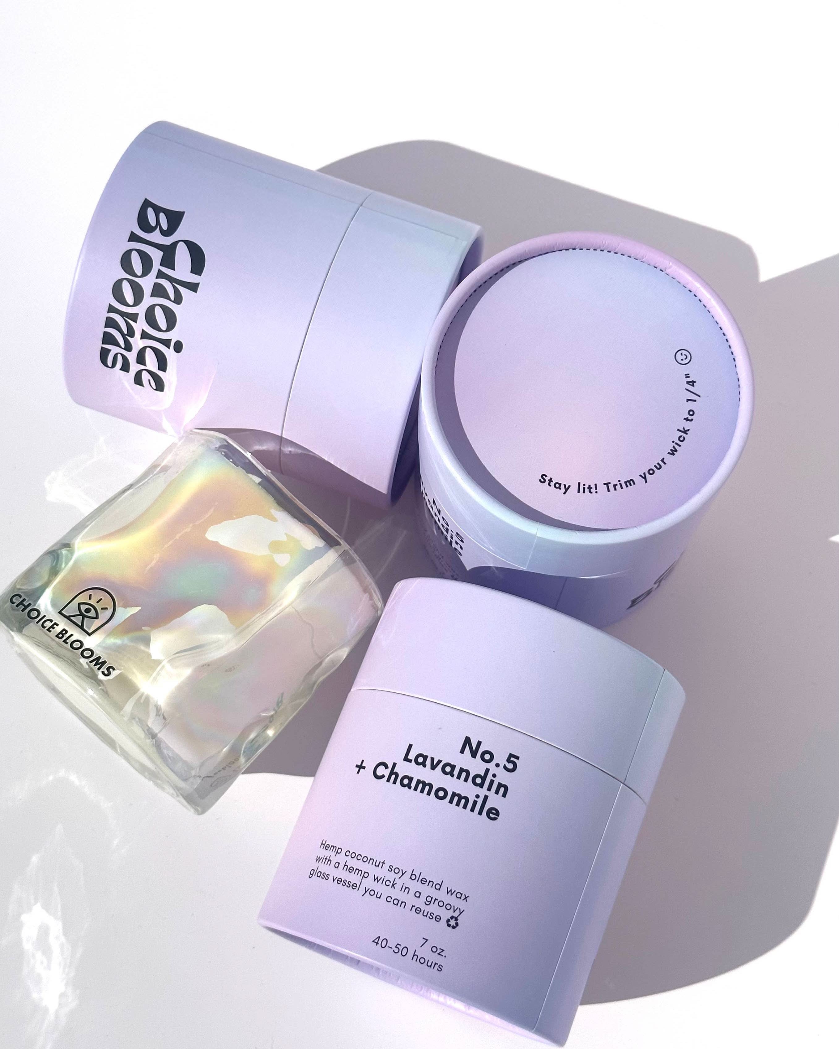 One candle in iridescent wavy glass cup laying on its side next to three purple packaging containers