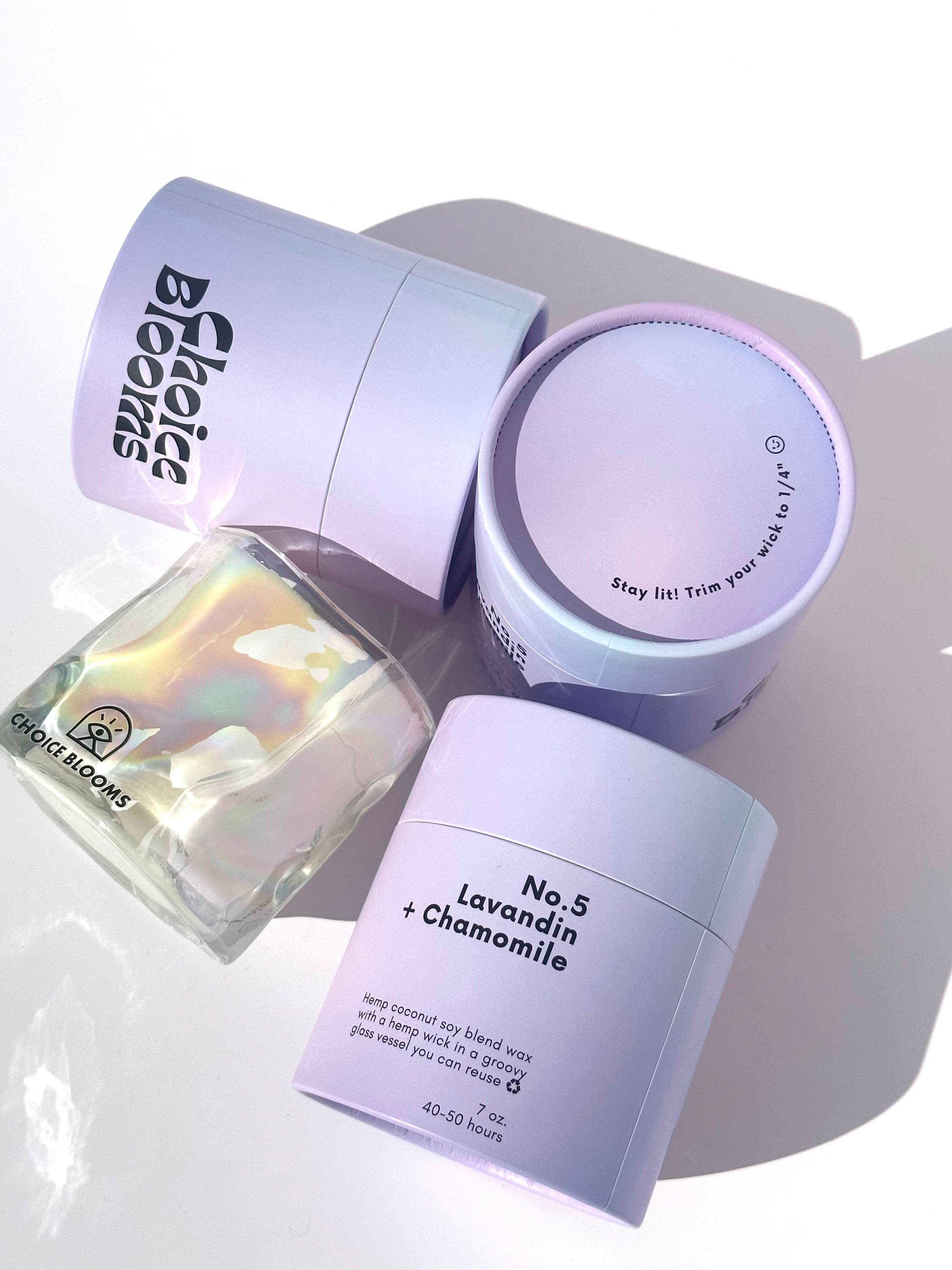 One candle in iridescent wavy glass cup laying on its side next to three purple packaging containers