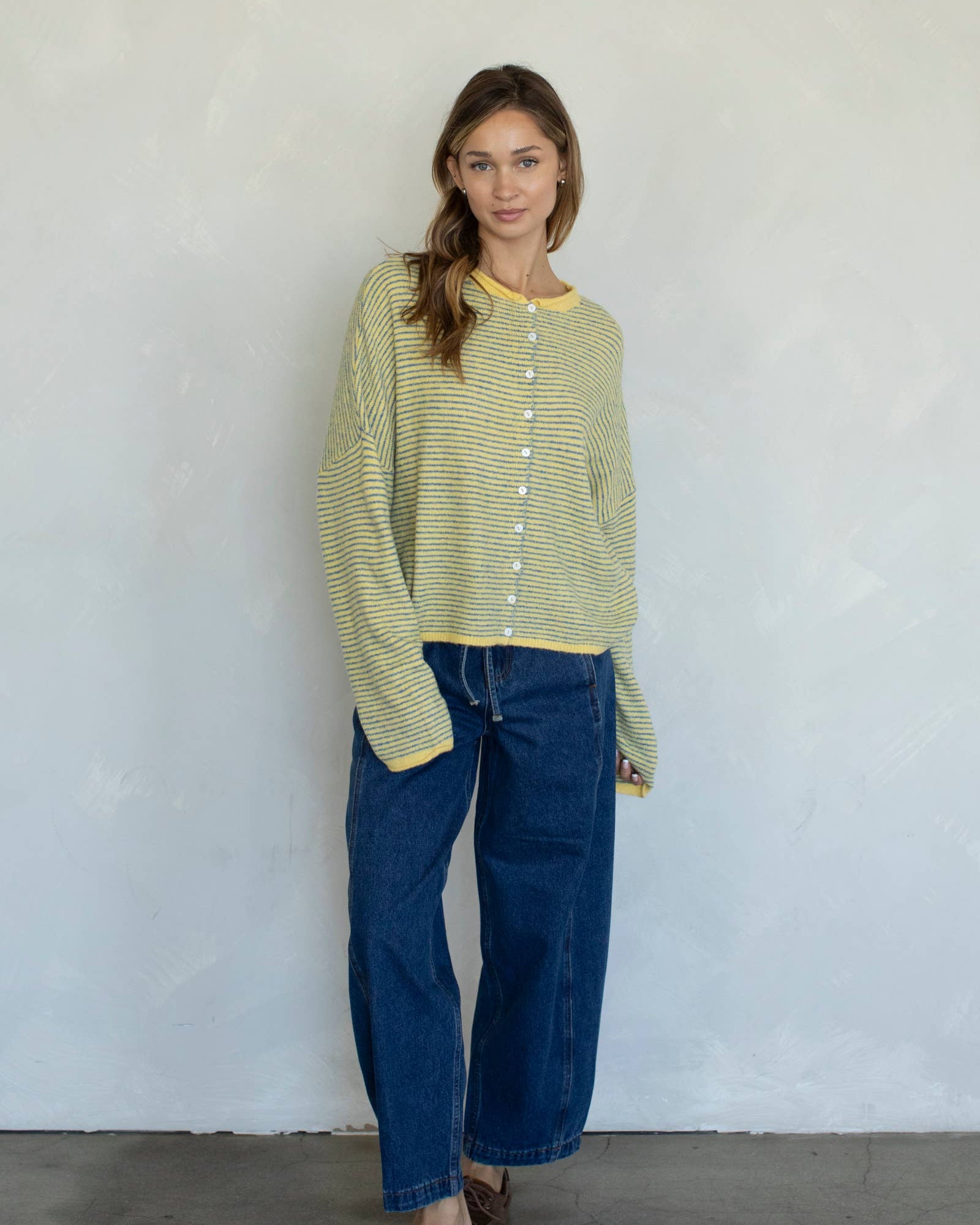 Woman wearing a green striped shirt and blue jeans standing against a white wall.