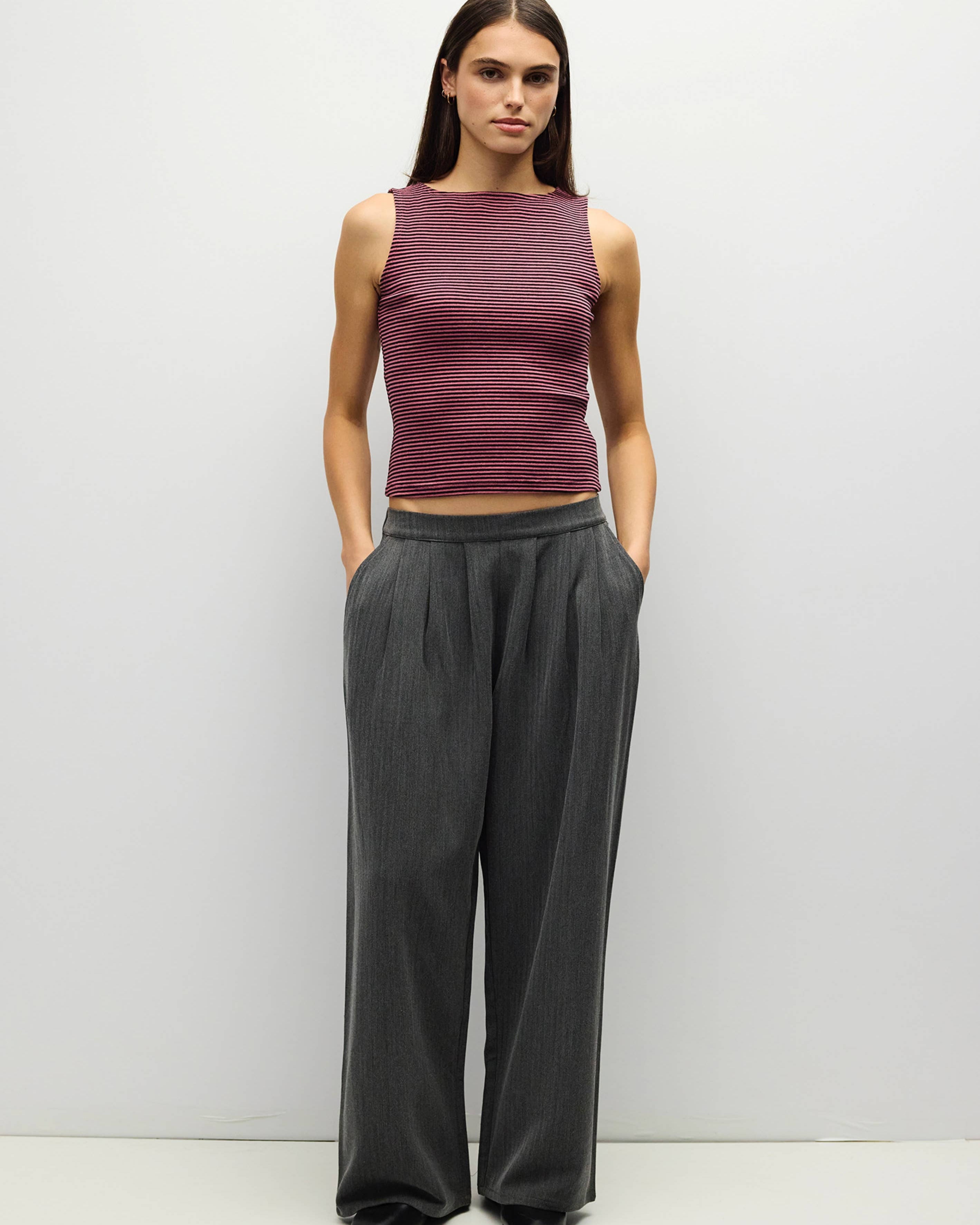 A woman stands against a plain light gray studio wall wearing a sleeveless heathered plum crop top and dark charcoal wide-leg trousers.