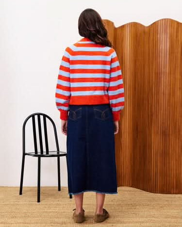 Person wearing a red and white striped sweater and blue skirt standing in a room with a black chair and wooden wall.