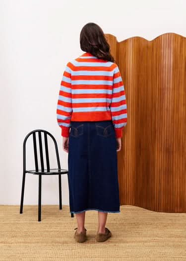 Person wearing a red and white striped sweater and blue skirt standing in a room with a black chair and wooden wall.