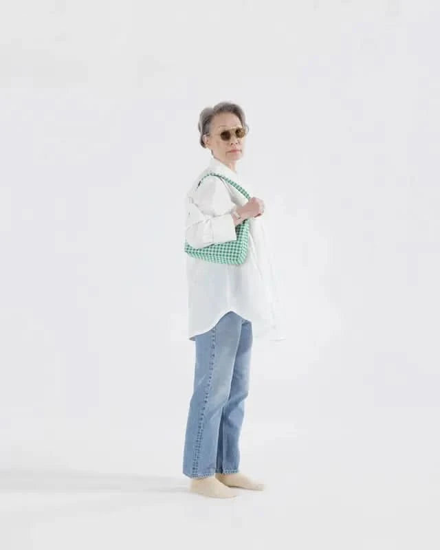 Model wearing white top and jeans holding BAGGU Green gingham nylon shoulder bag sitting against white background
