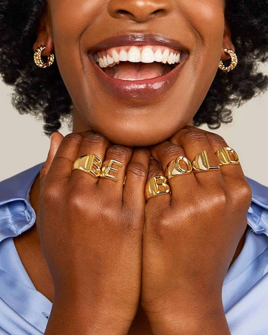 Smiling woman wearing gold letter rings in the words BE BOLD