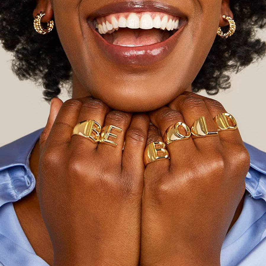 Smiling woman wearing gold letter rings in the words BE BOLD