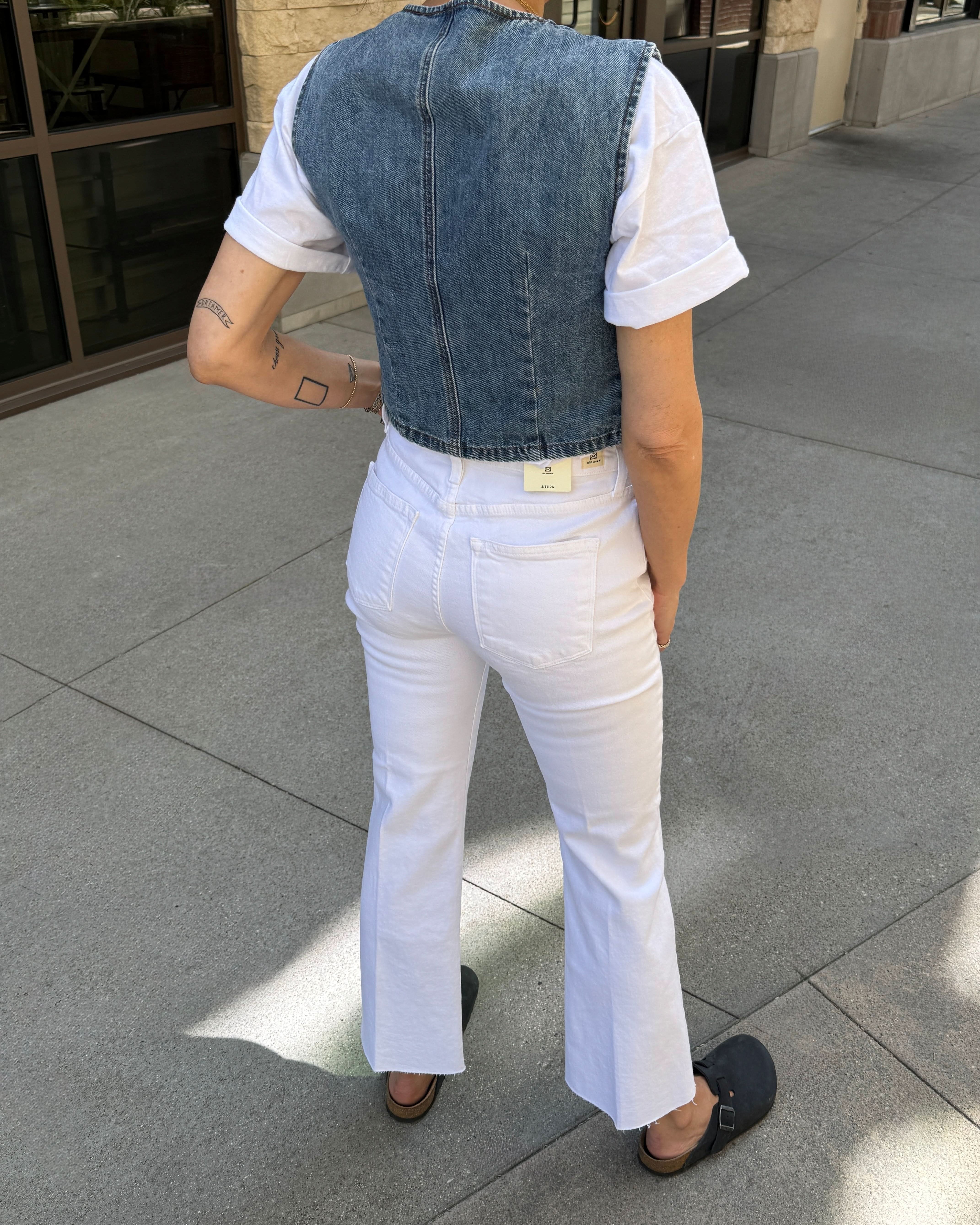 Person wearing a denim vest over a white shirt and pants on a sidewalk.