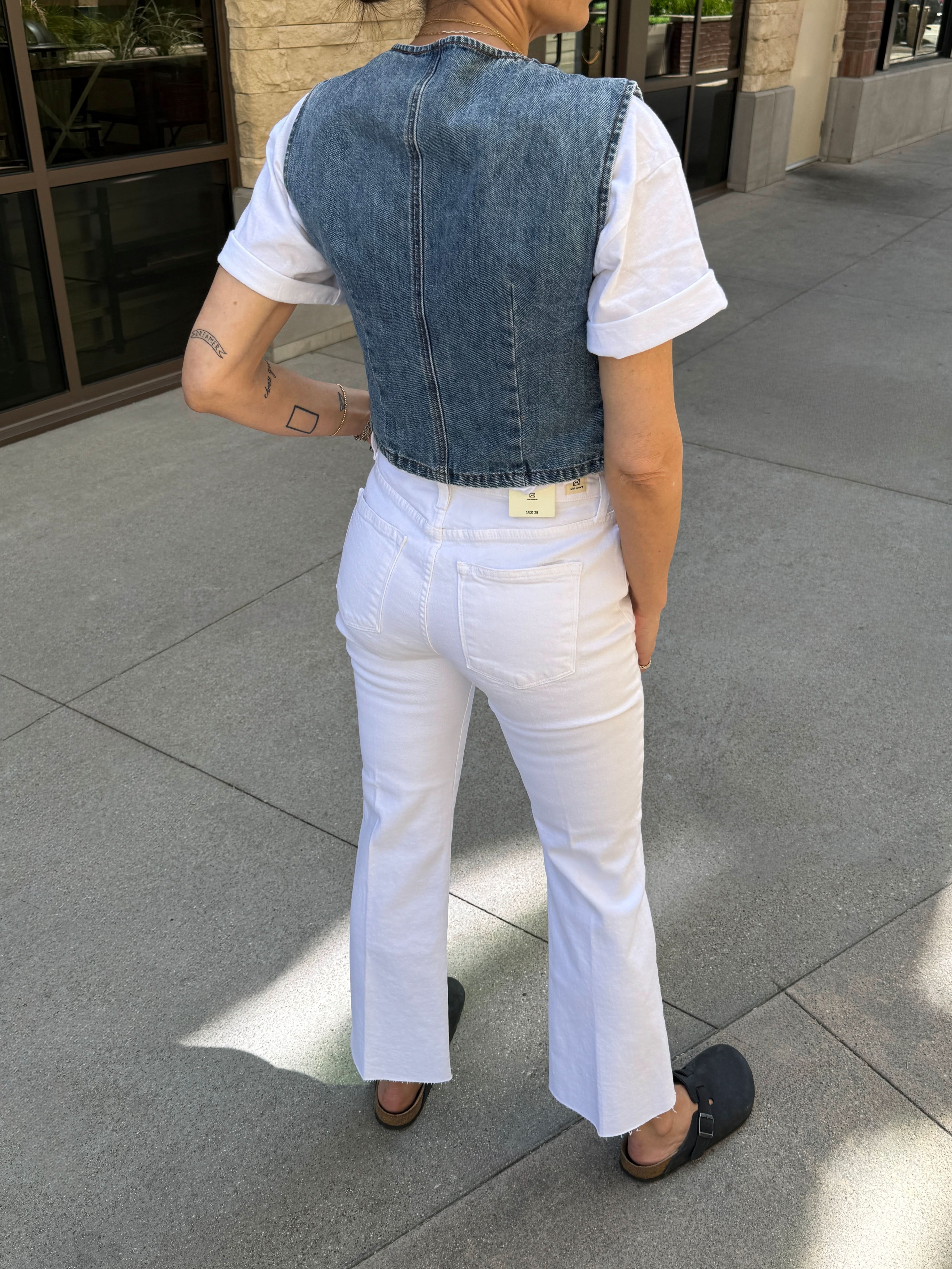Person wearing a denim vest over a white shirt and pants on a sidewalk.