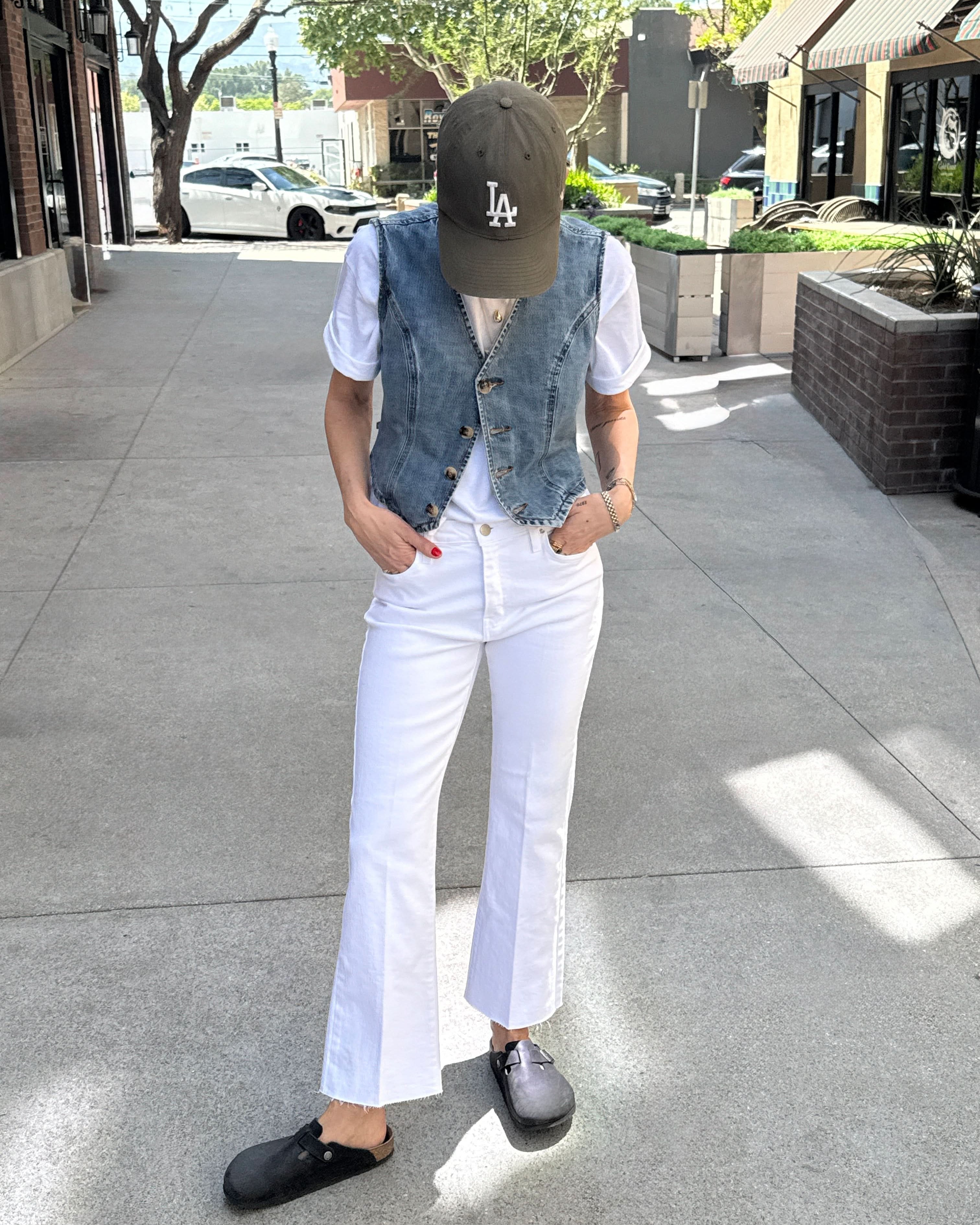 Person wearing a denim vest, white shirt, white pants, and black shoes on a sidewalk.