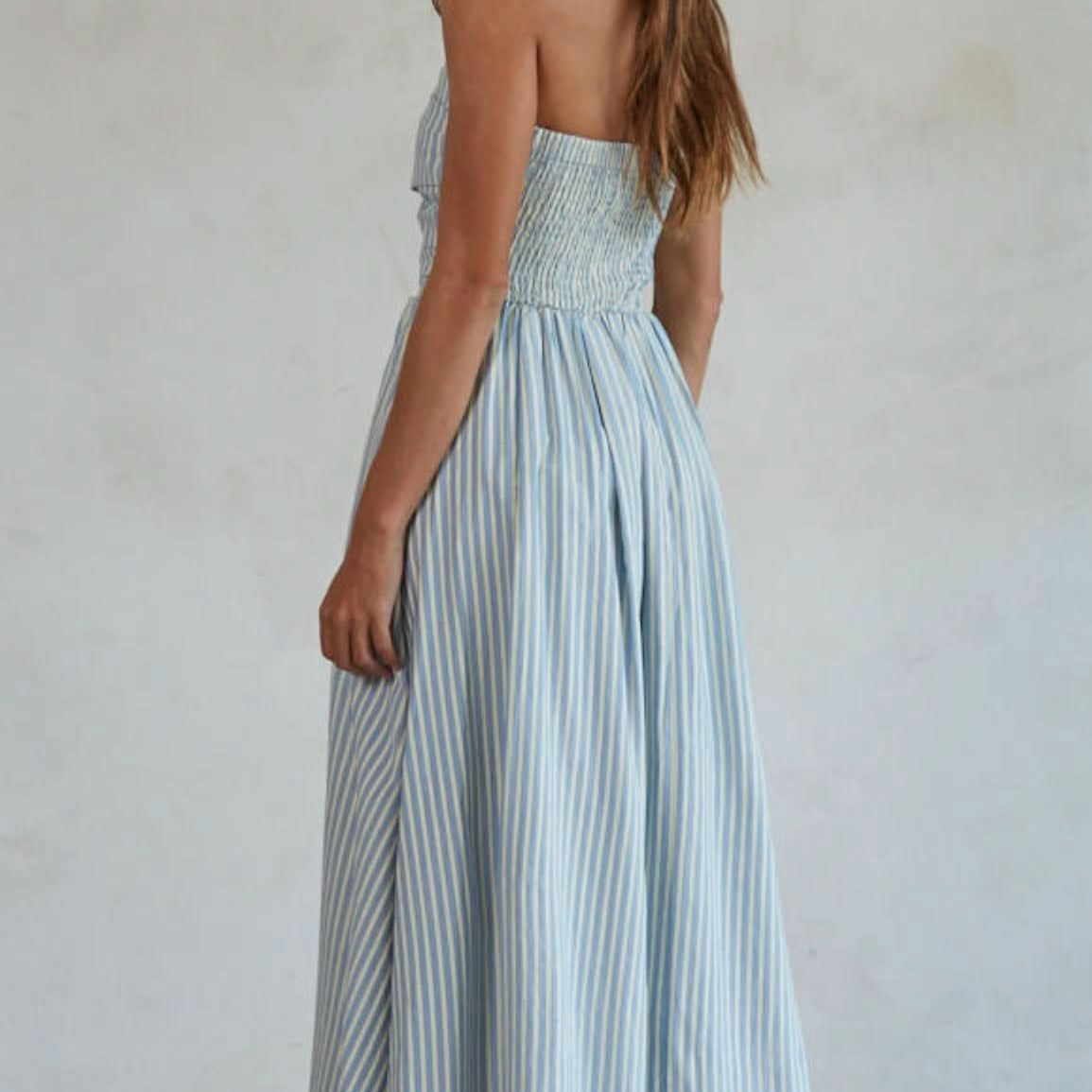Woman wearing white and blue vertically striped, sleeveless maxi dress standing against a blank wall