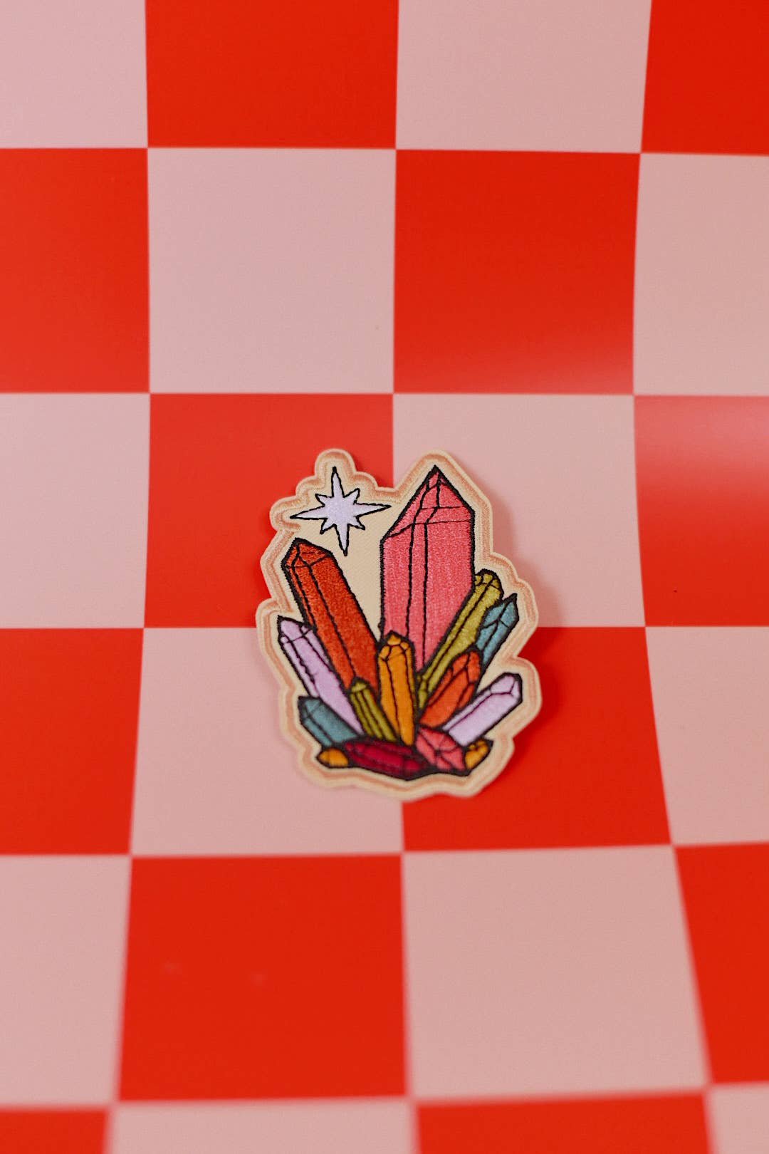 A die-cut enamel pin featuring a colorful cluster of crystals, displayed on a red and white checkered background.