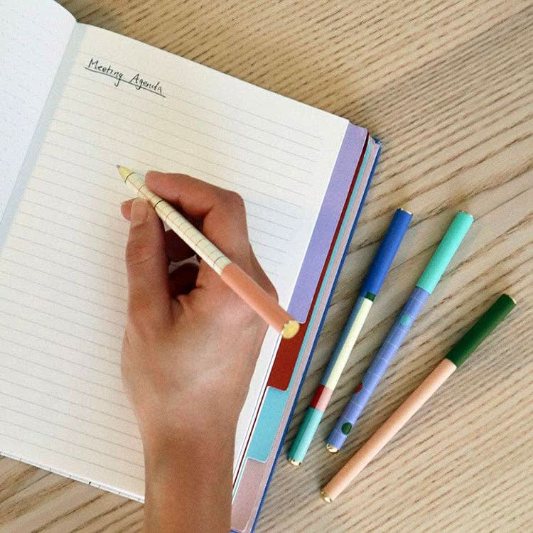 Four pens and a notebook opened and someone using one of the pens to write in notebook